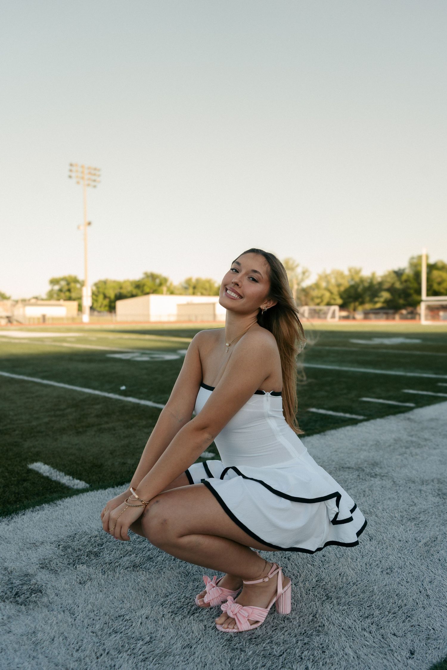Smiling young woman in white dress with pink heels crouching on green football field at sunset.