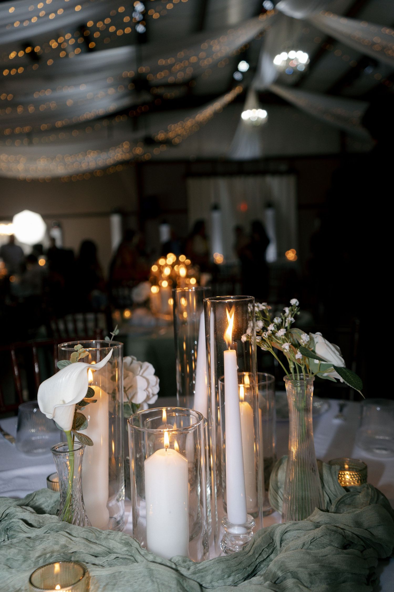 Elegant wedding reception centerpiece with white pillar candles in glass vases and white flowers on sage green table runner.