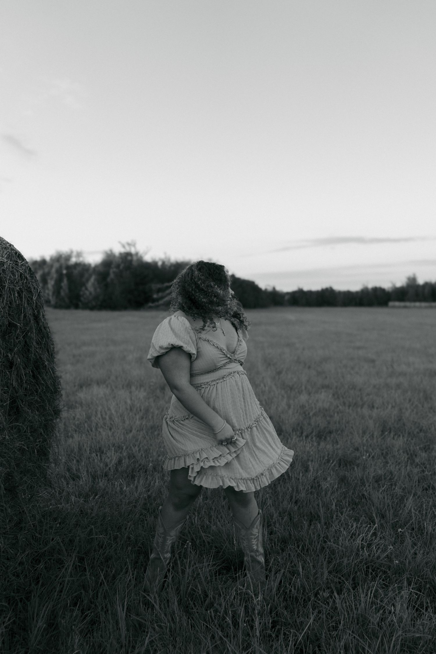 Black and white photo of young woman in ruffled dress standing beside a hay bale in an open field.