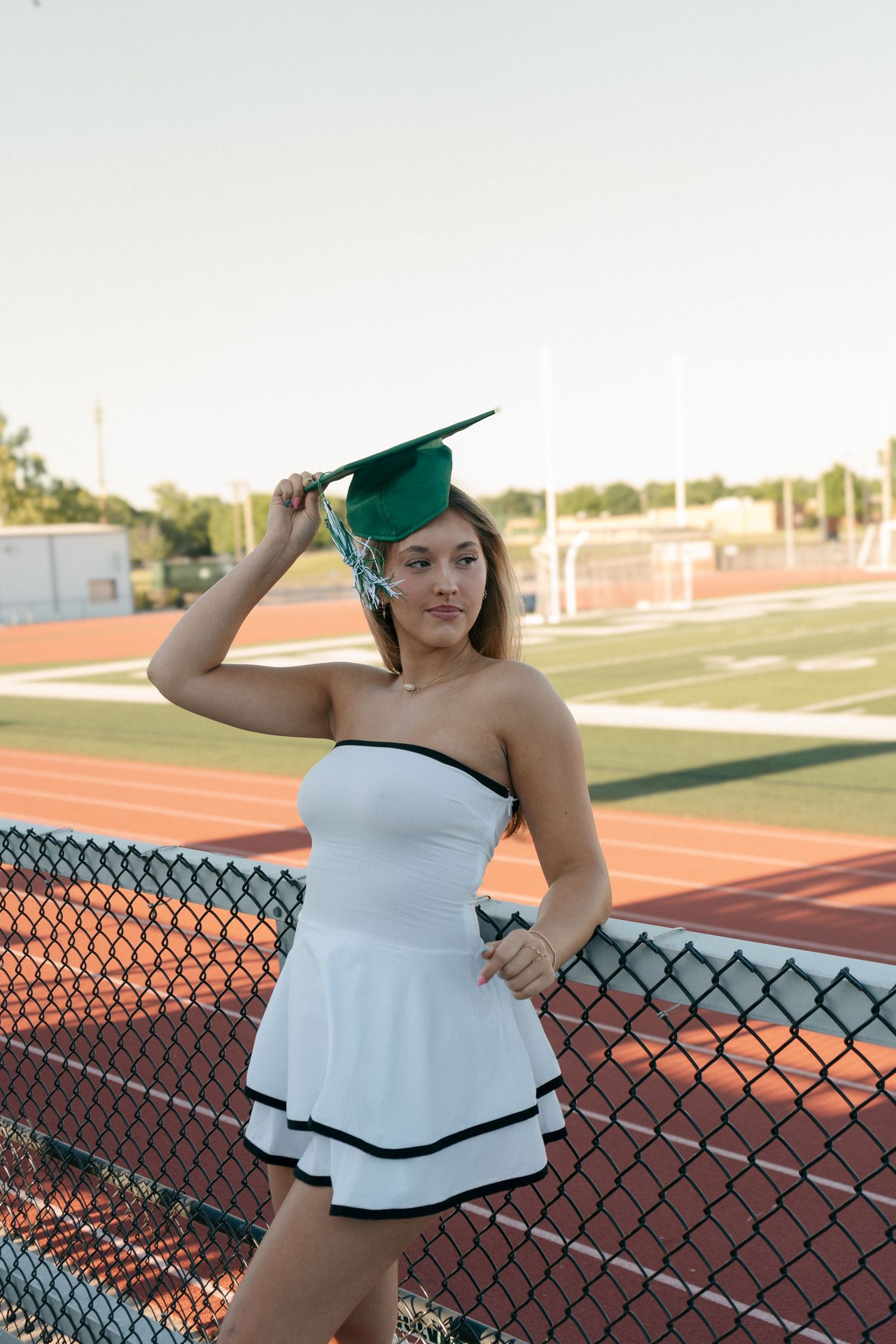 Graduate in white strapless dress holds green mortarboard at school track.