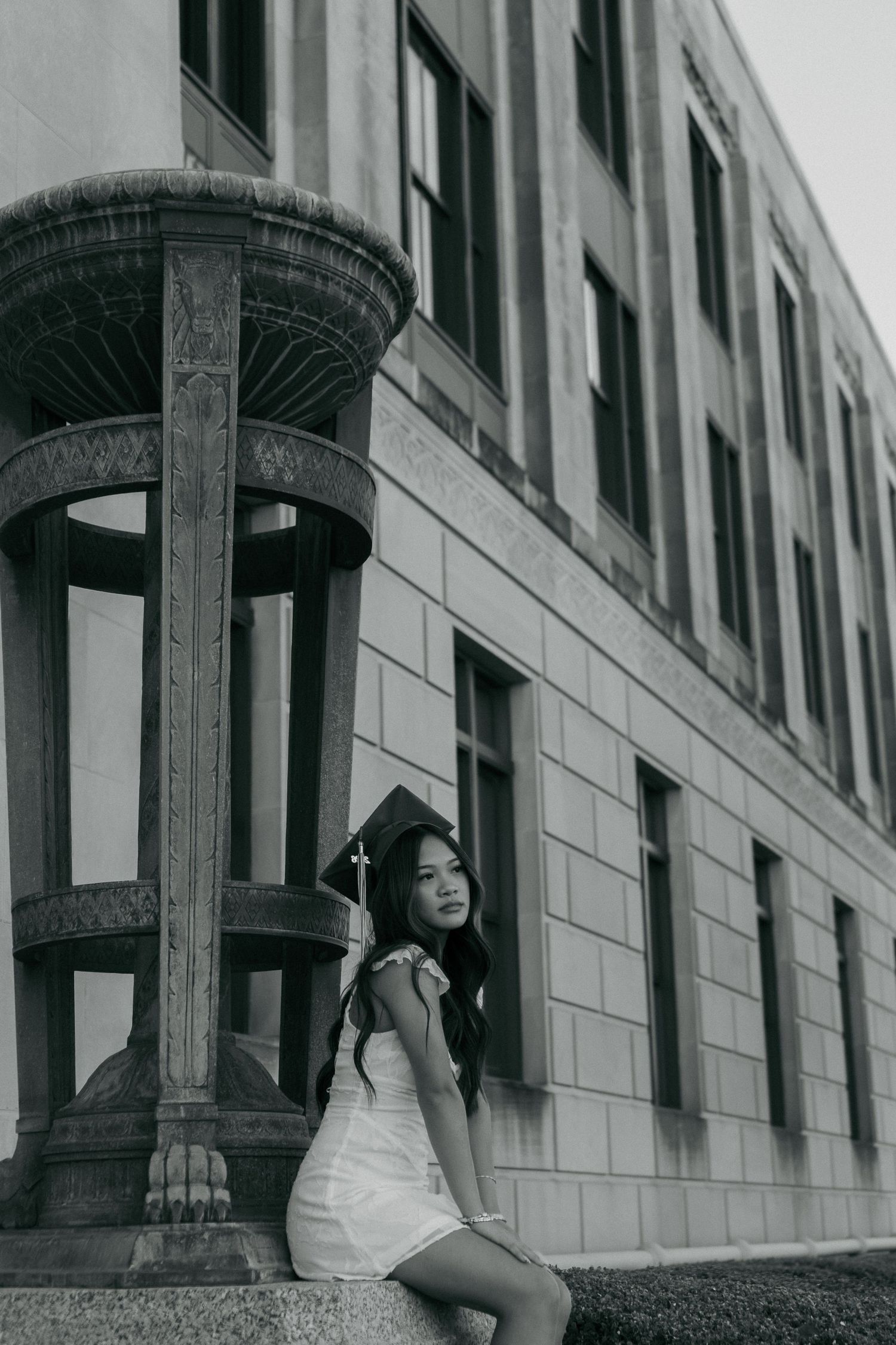 Black and white senior portrait of young woman in white dress sitting by ornate stone column outside.