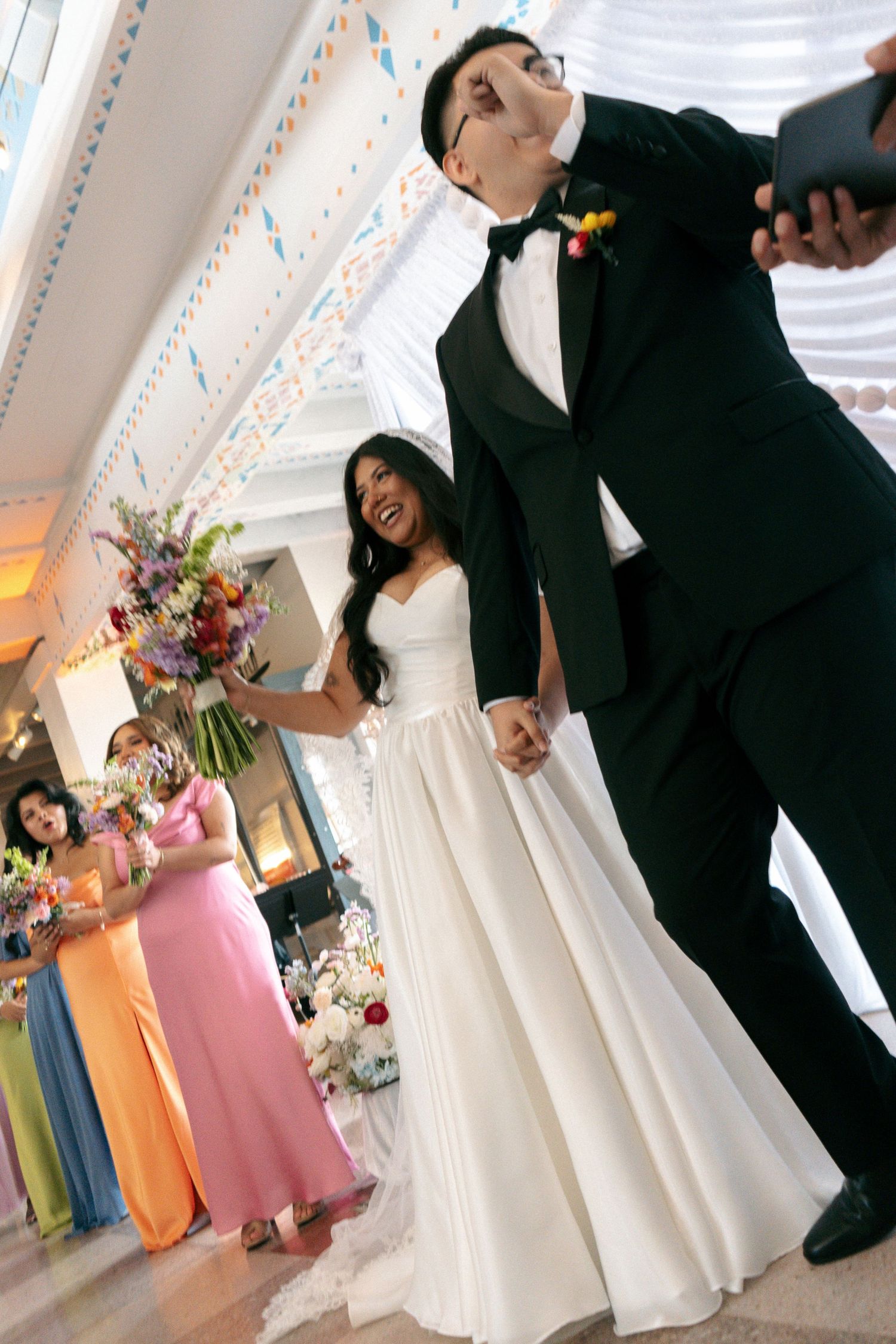 Bride and groom hold hands at wedding reception, bridesmaids in colorful dresses stand behind them.