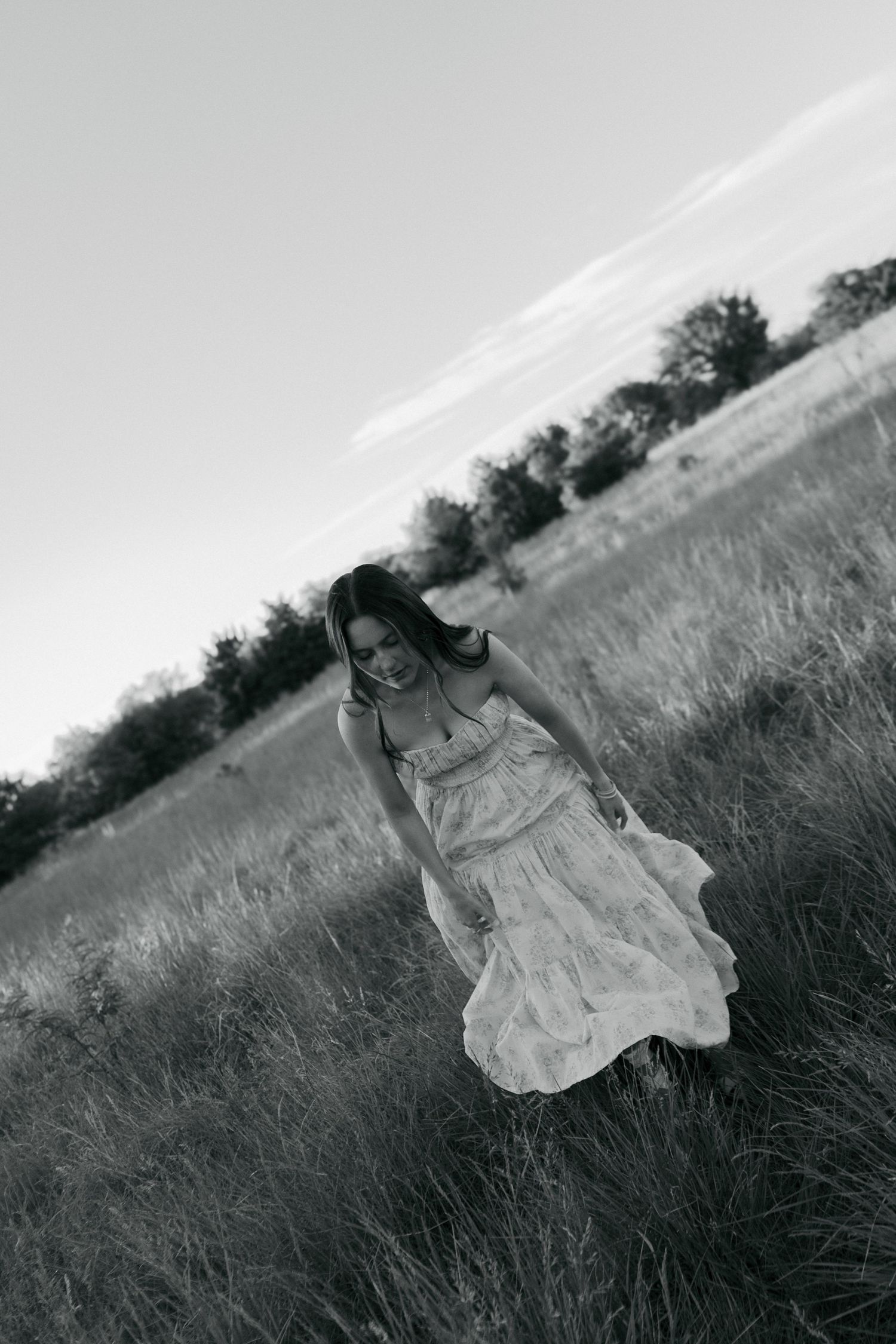 Black and white photo of a woman in a flowing dress walking through a grassy field with trees in the background.