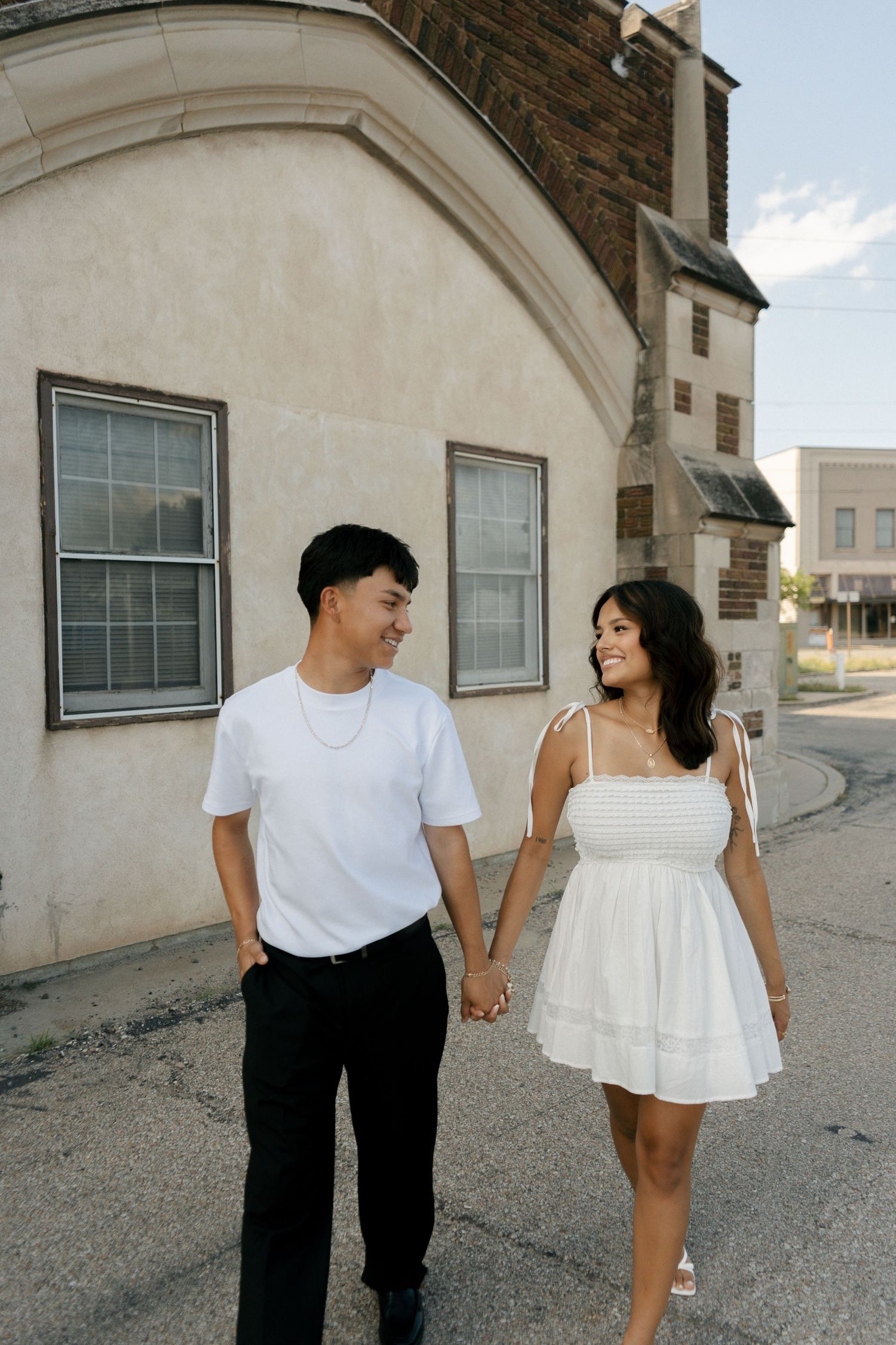 Smiling couple holding hands walking past a vintage building on a sunny day.