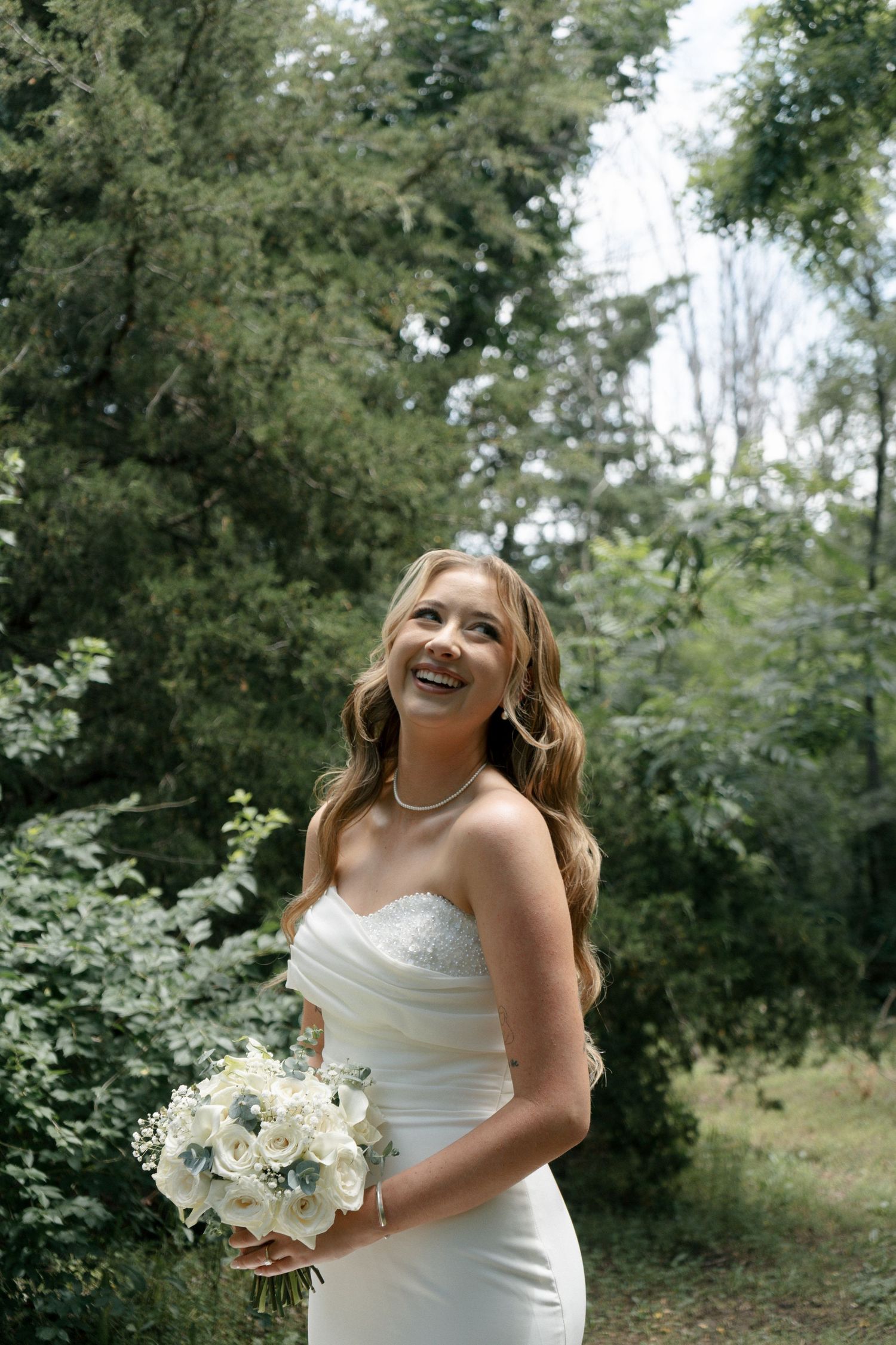 Smiling bride in strapless white gown holds white floral bouquet outdoors surrounded by lush green trees.