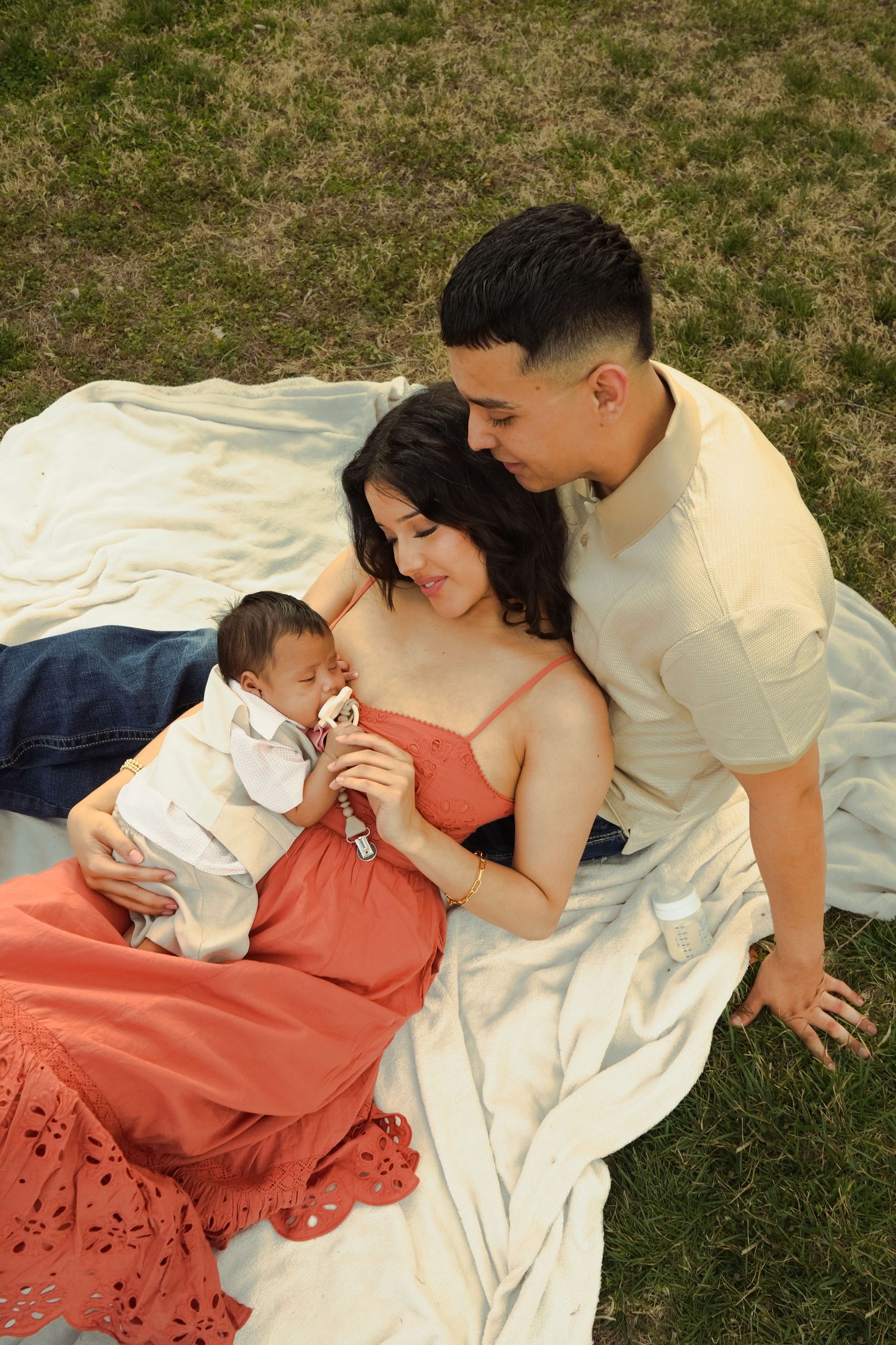 Young couple sitting on blanket in grass lovingly gazing at newborn baby in mother's arms.