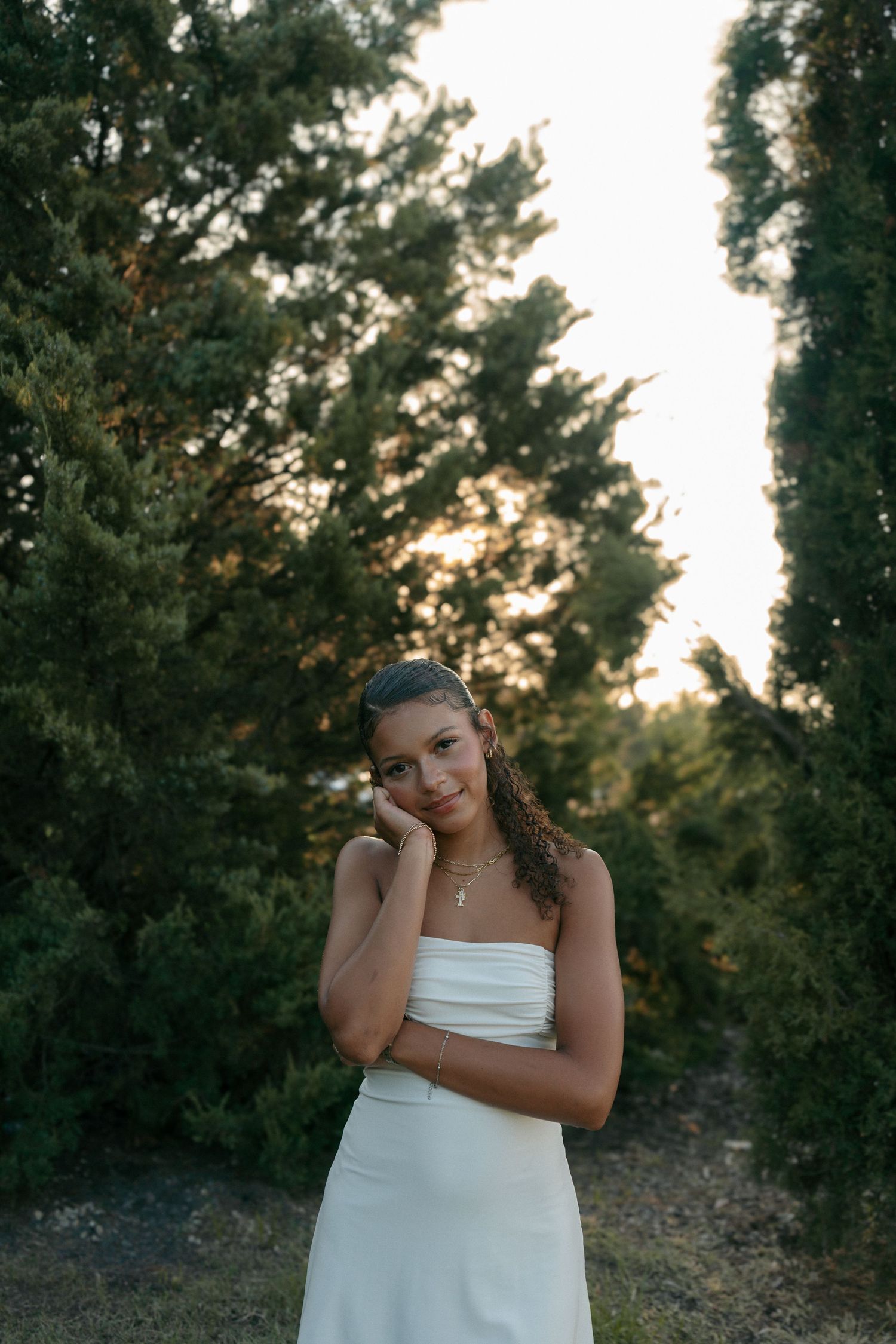 Young woman in white strapless dress posing thoughtfully between tall cypress trees at golden hour sunset.