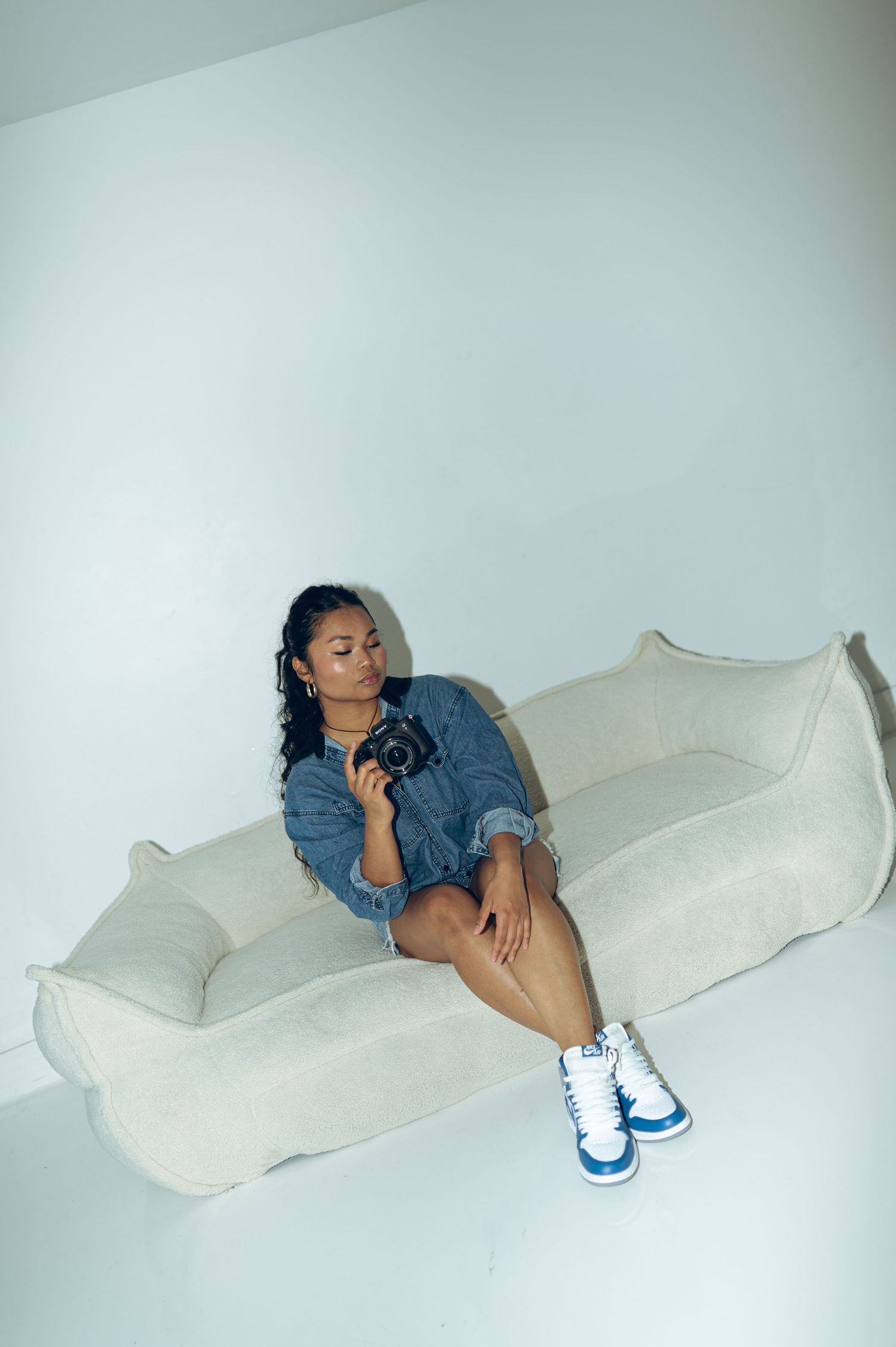 Young woman in denim jacket sitting on a beige sofa holding a vintage camera in a white studio.