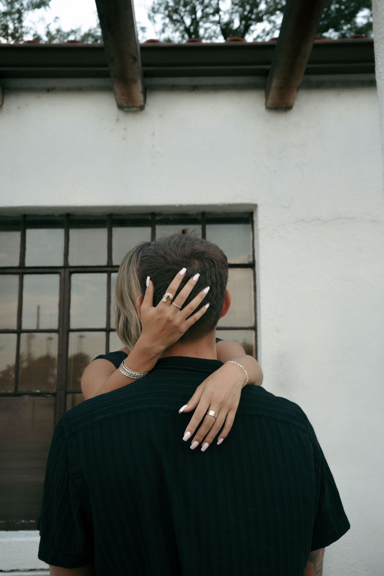 Woman with manicured nails and bracelets embracing man from behind near white wall.