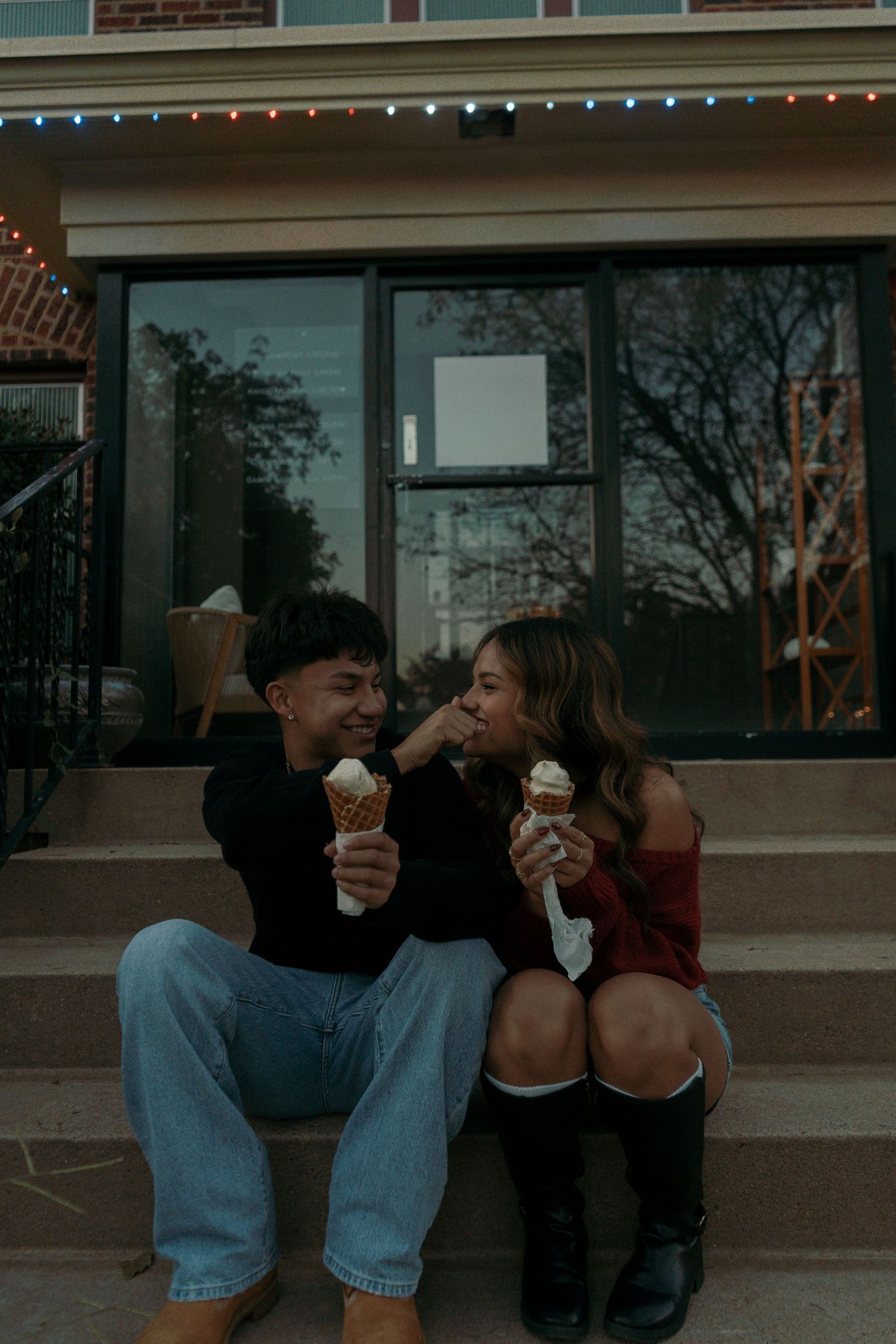 Playful couple laughing and sharing ice cream cones on outdoor steps with string lights above.