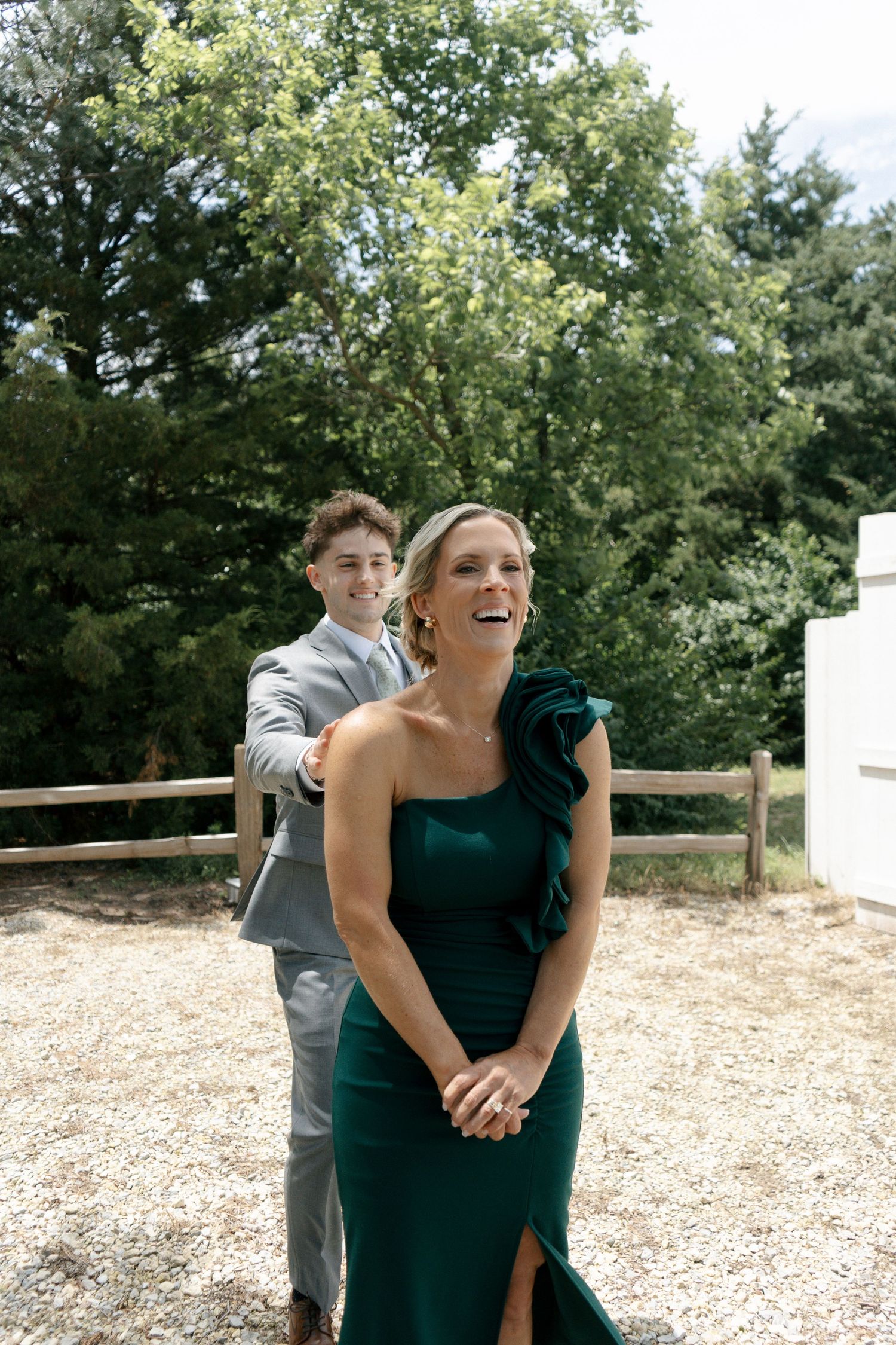 A woman in an emerald green gown laughs joyfully as a young man in a gray suit stands behind her outdoors.
