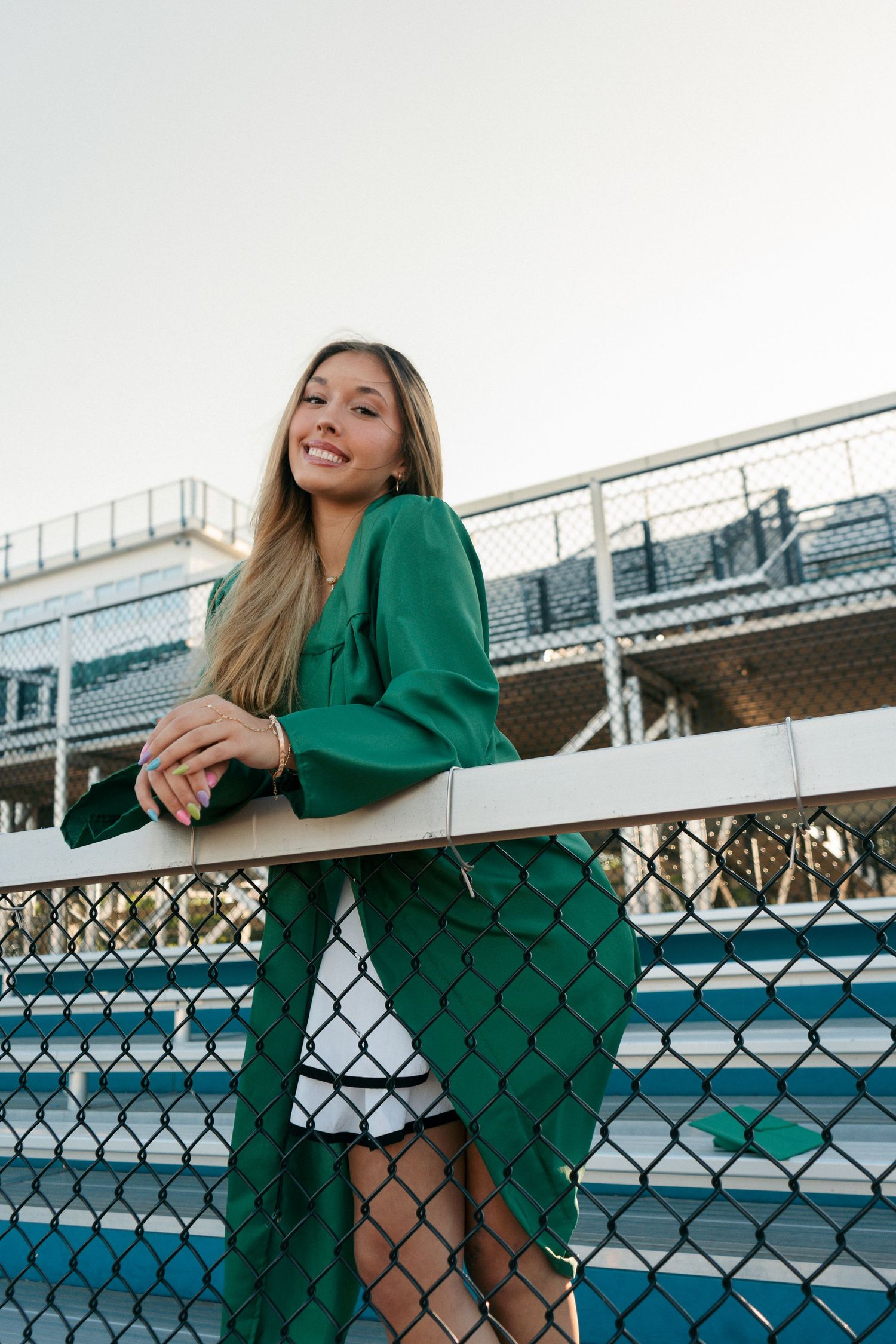 Happy graduate in green gown leans on white railing at school stadium.