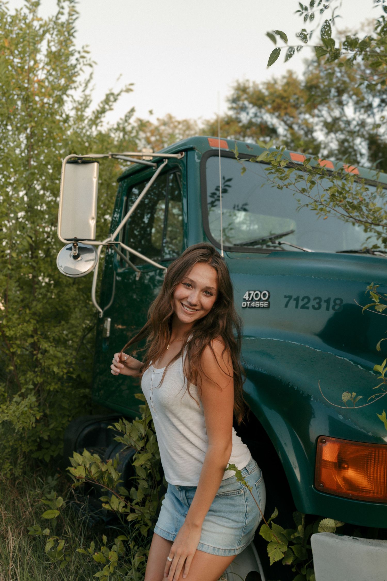 Smiling young woman in white tank and denim shorts leaning against a vintage green truck outdoors.