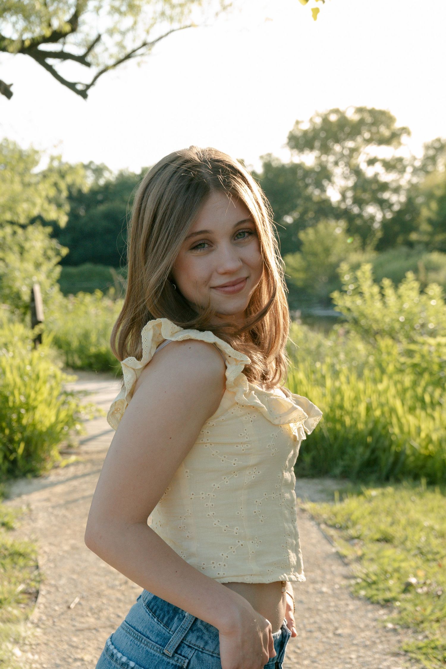 Young blonde girl in yellow ruffled top and jeans smiling on a sunny garden path.