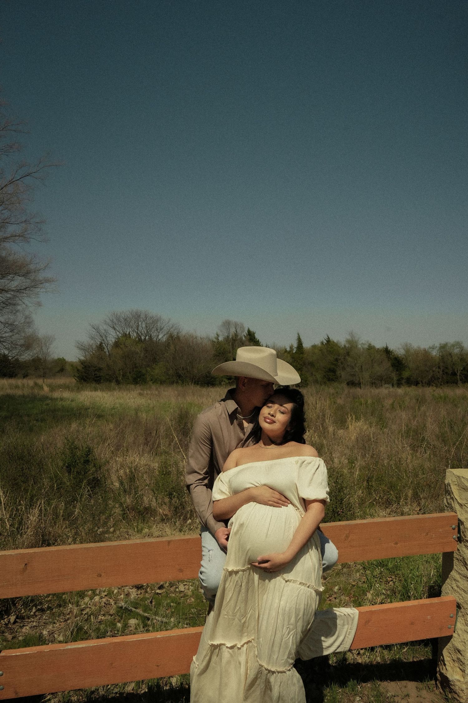Couple poses outdoors during maternity shoot near wooden fence, man in cowboy hat kisses pregnant womans head.