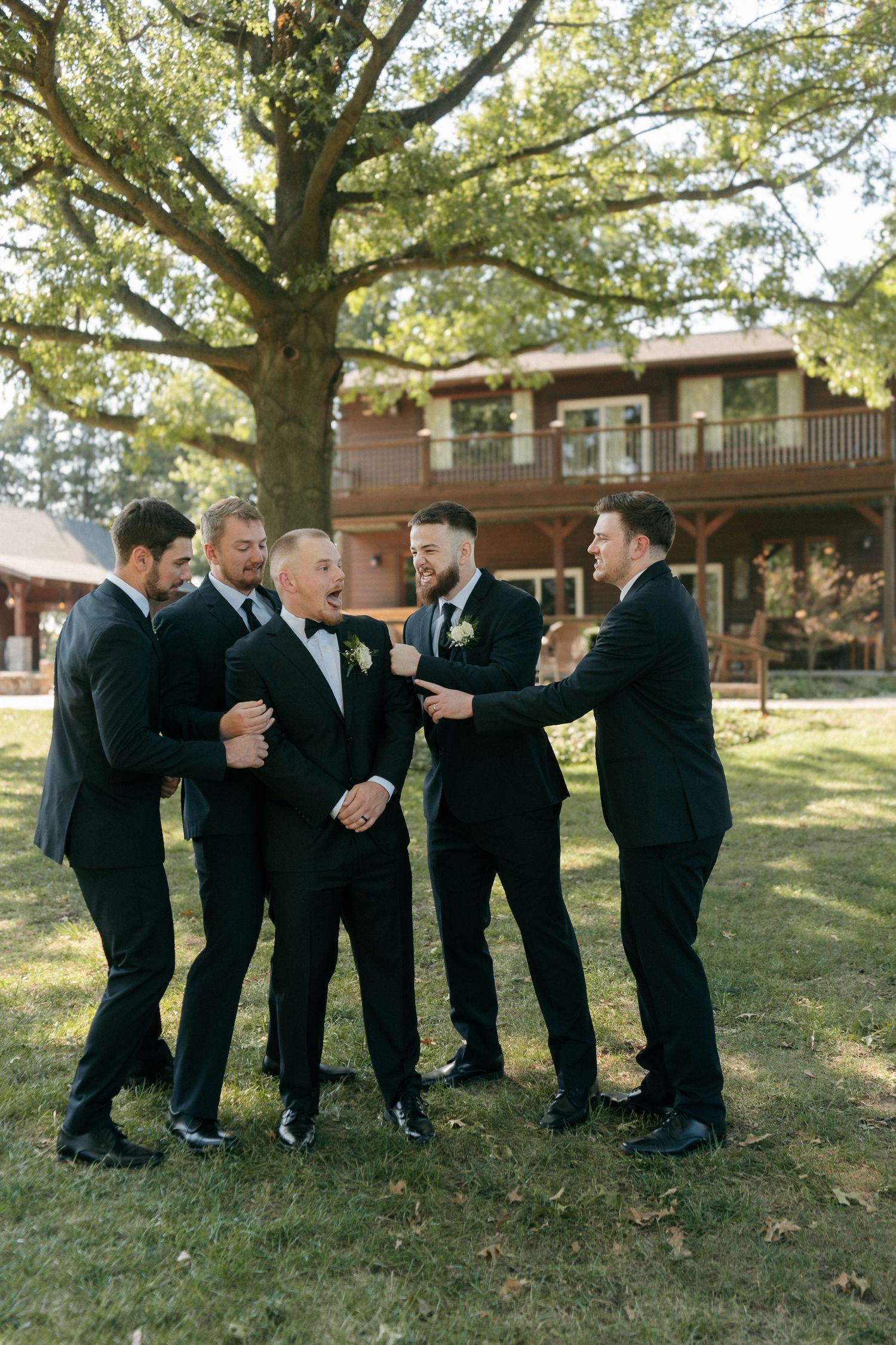 Groom and groomsmen in black tuxedos laughing together outdoors near rustic lodge wedding venue.