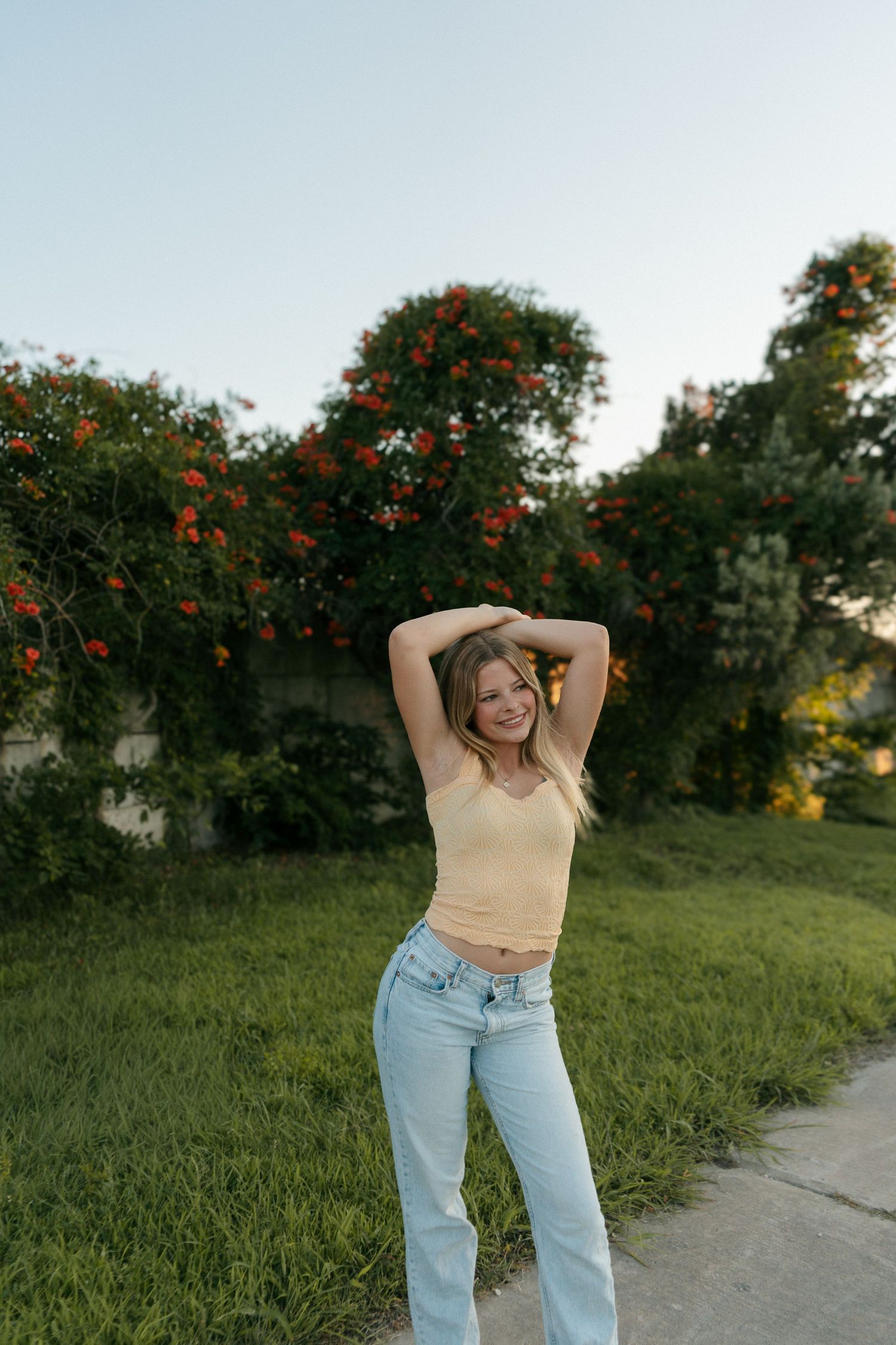 Young woman in yellow crop top and light blue jeans posing outdoors with hands raised, smiling near flowering bushes.