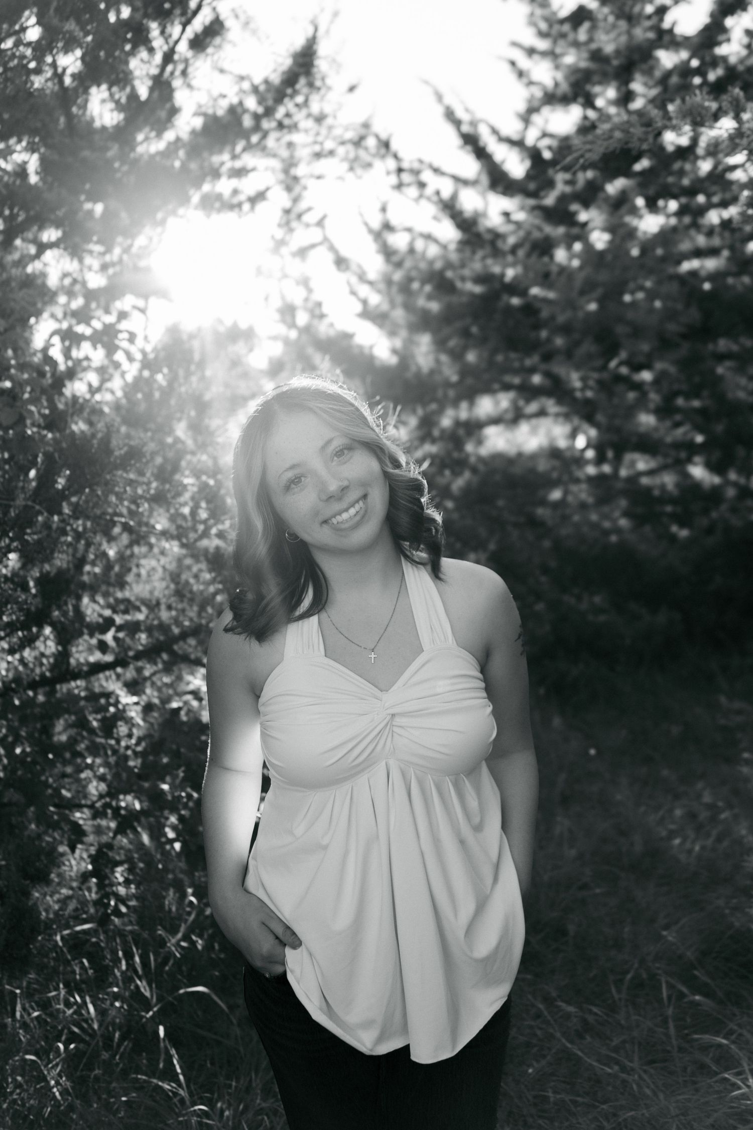 Black and white portrait of a smiling woman in a halter top standing outdoors with sunlight filtering through trees.