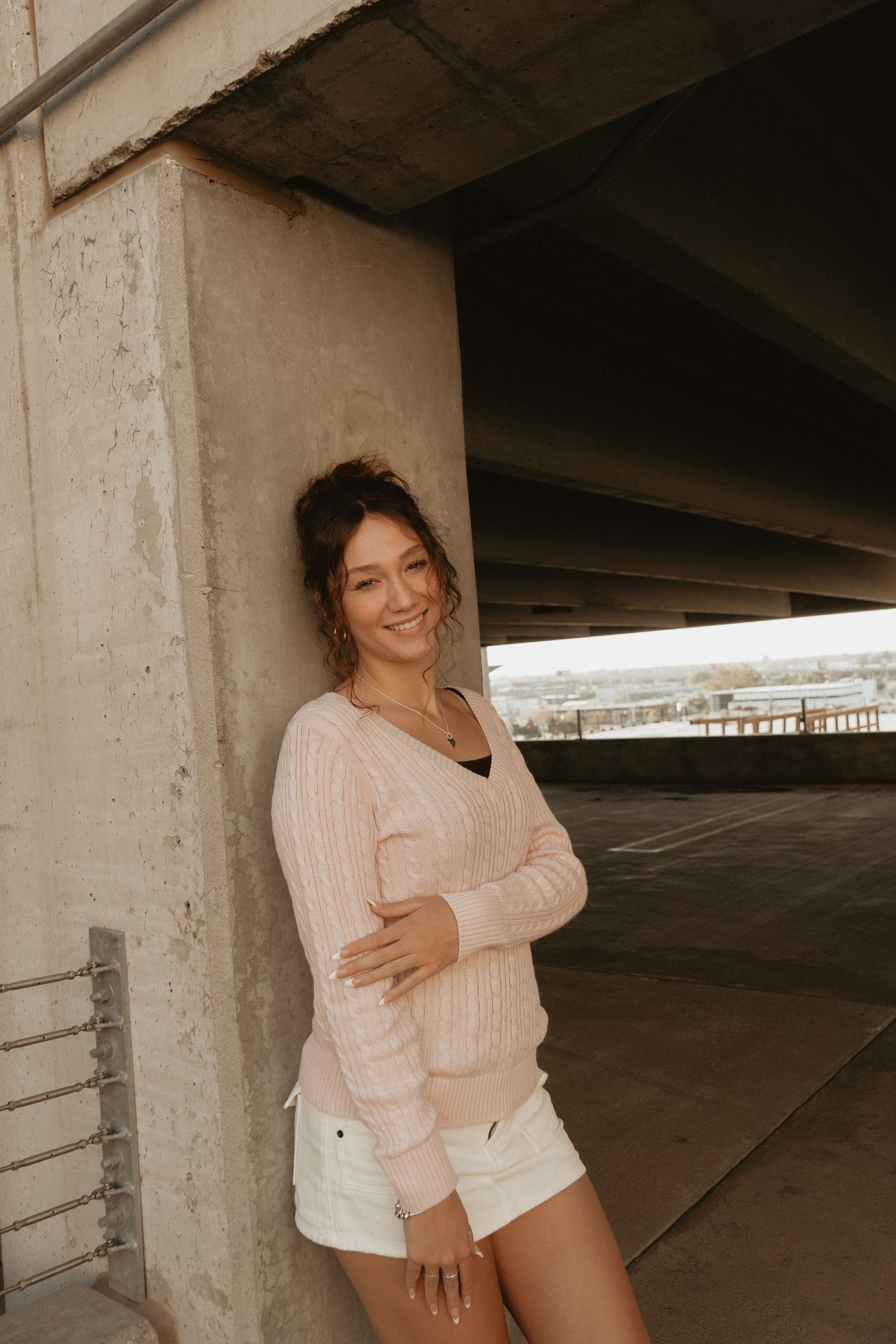 A smiling young woman in a pink sweater and white shorts leans against a concrete pillar outdoors.