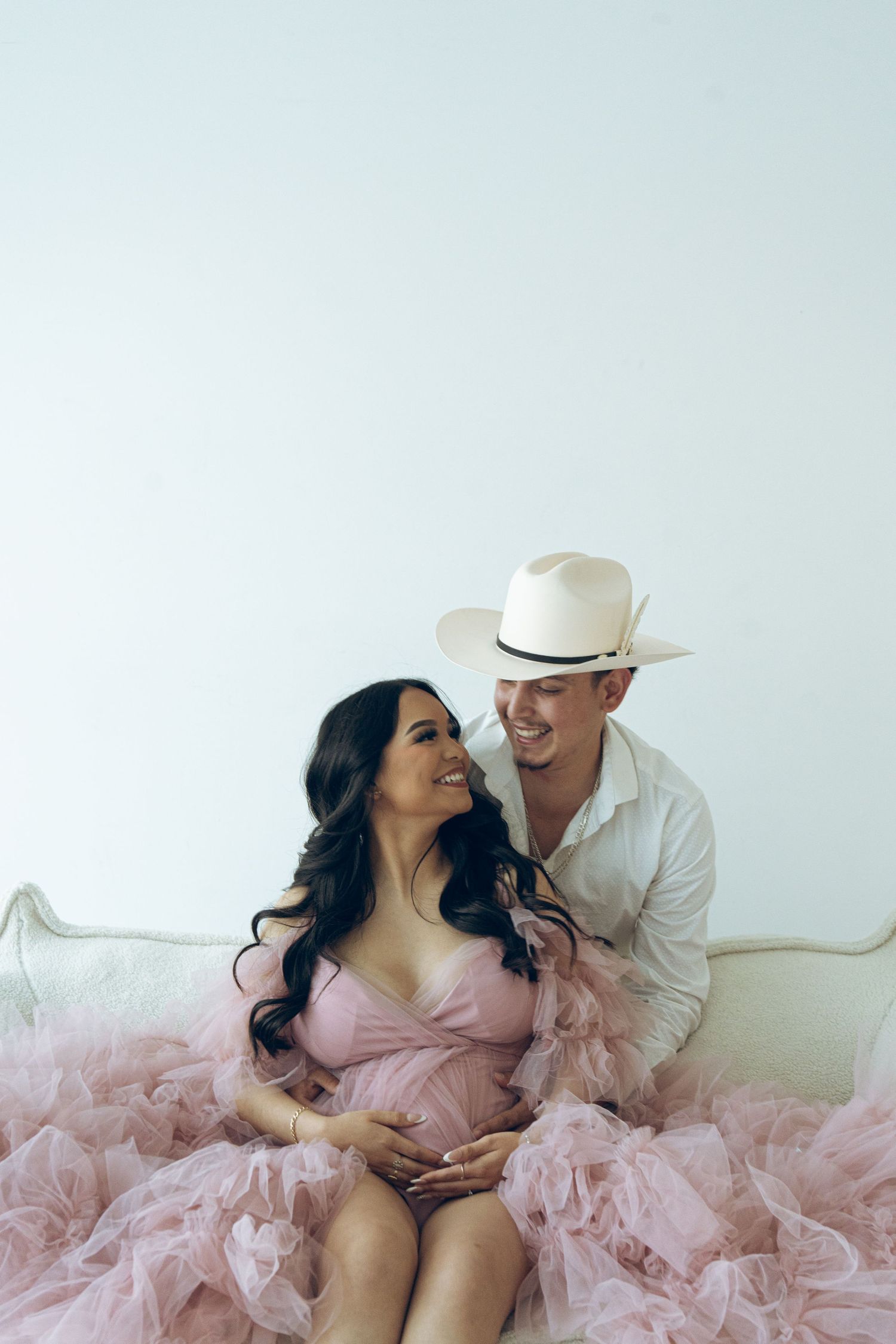 Pregnant woman in pink tulle dress sitting with man in cowboy hat on couch.