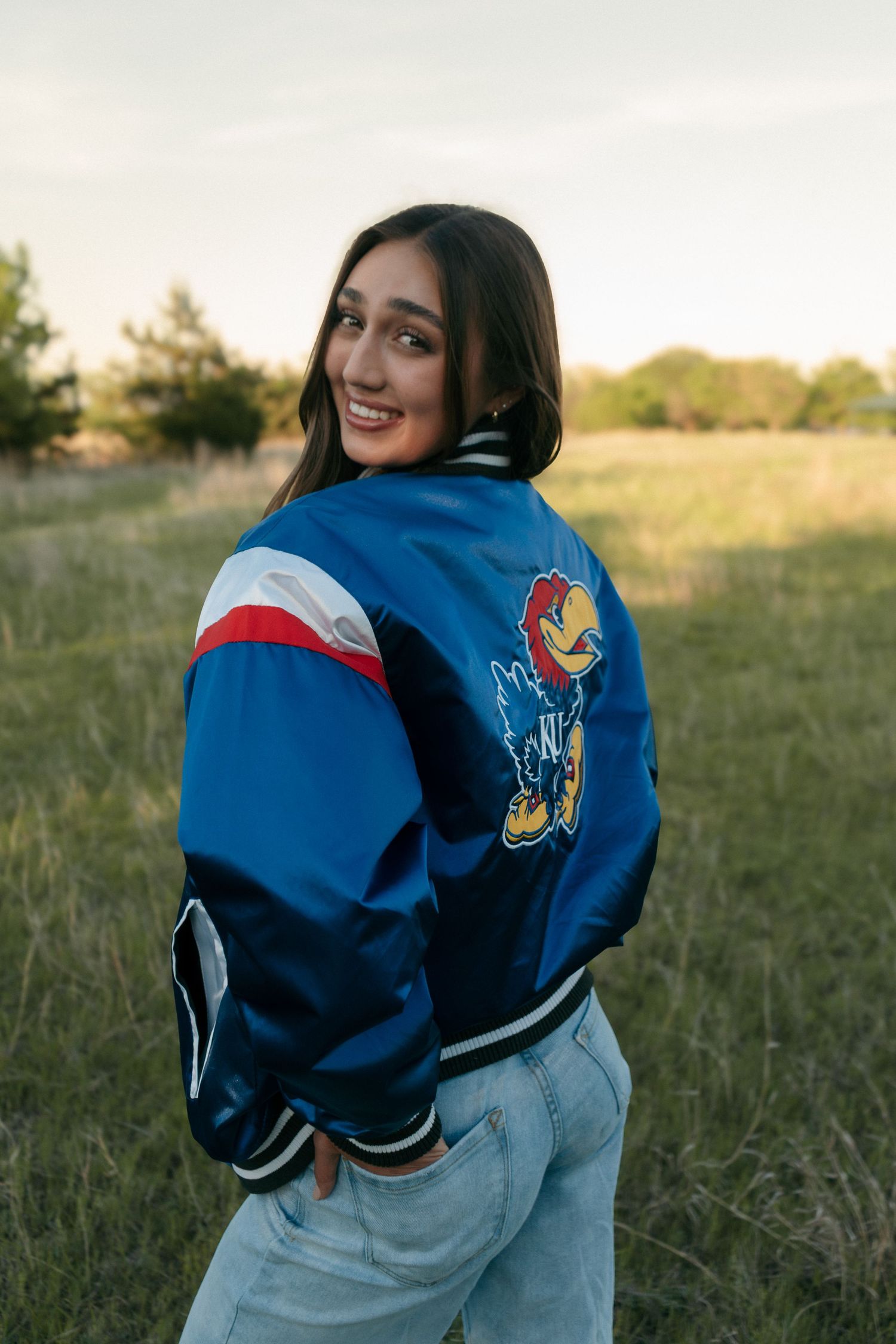Smiling young woman wearing Kansas Jayhawks blue varsity jacket posing in golden hour field.