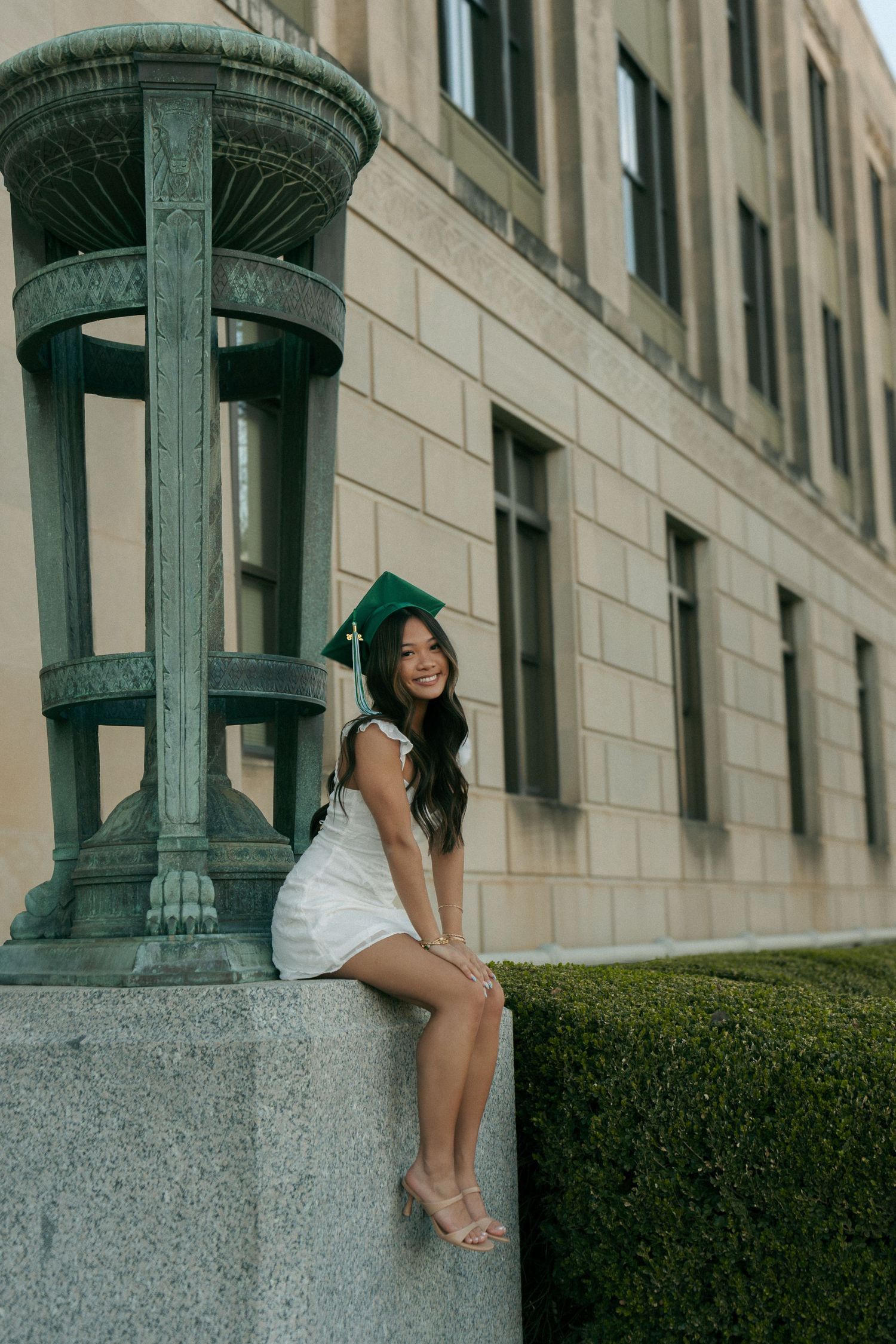 Smiling graduate in white dress and green cap poses beside a decorative stone column on campus.
