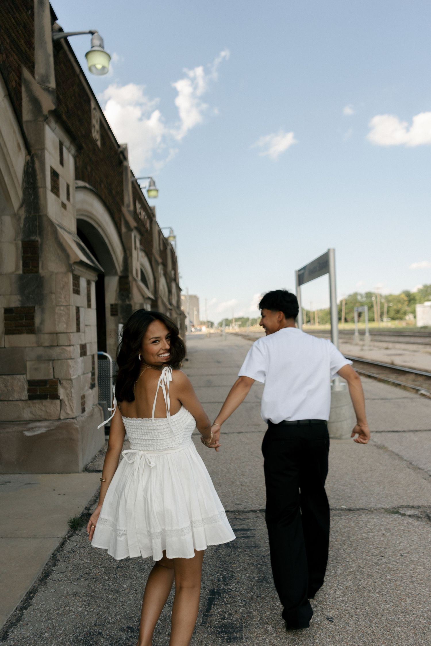 A couple holds hands walking along a train station platform, woman in white dress smiling back at camera.