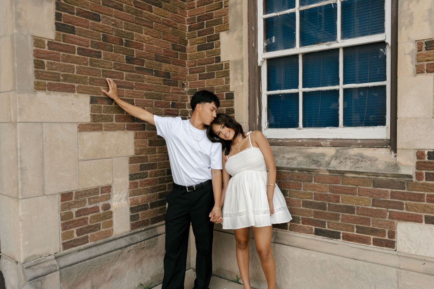 Couple posing romantically against a brick wall, man in white tee and black pants, woman in white mini dress.