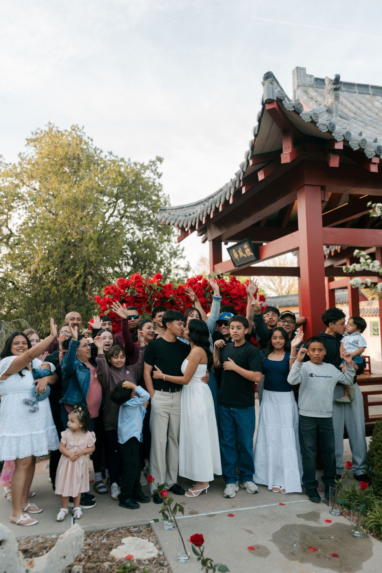 Large group of smiling people gathered under red floral arch at a traditional Chinese garden pavilion.