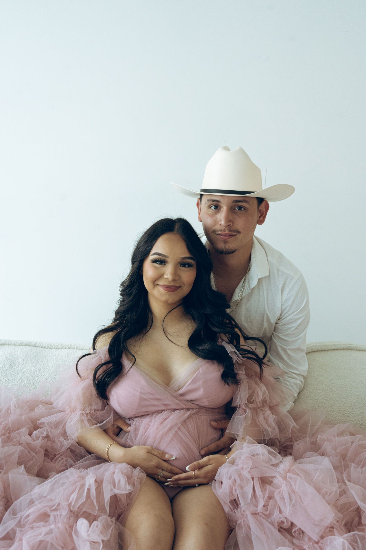 Smiling couple poses together during maternity shoot, woman in pink tulle gown and man in white cowboy hat.