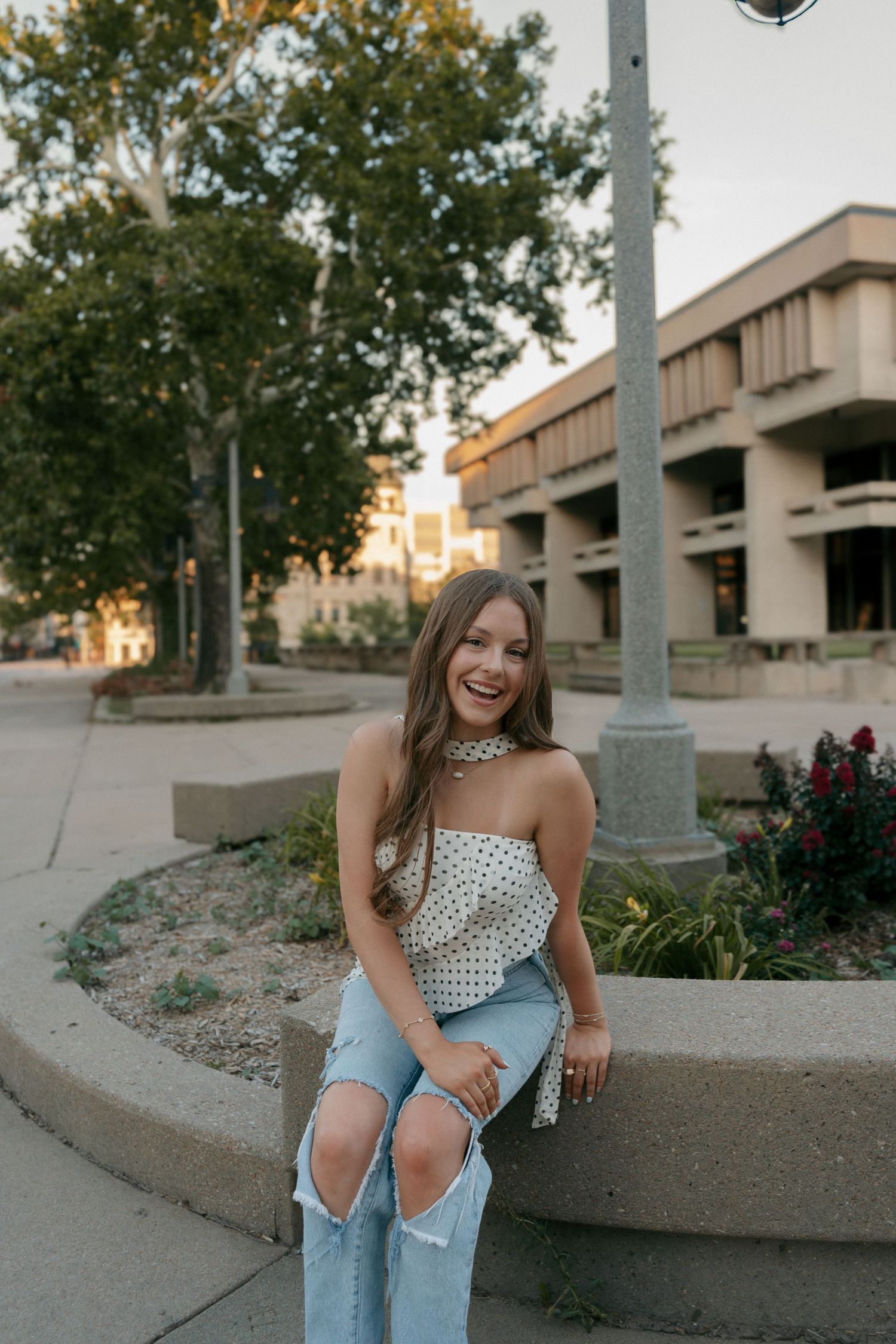 Laughing young woman in polka dot top and ripped jeans sits on a planter at golden hour downtown.