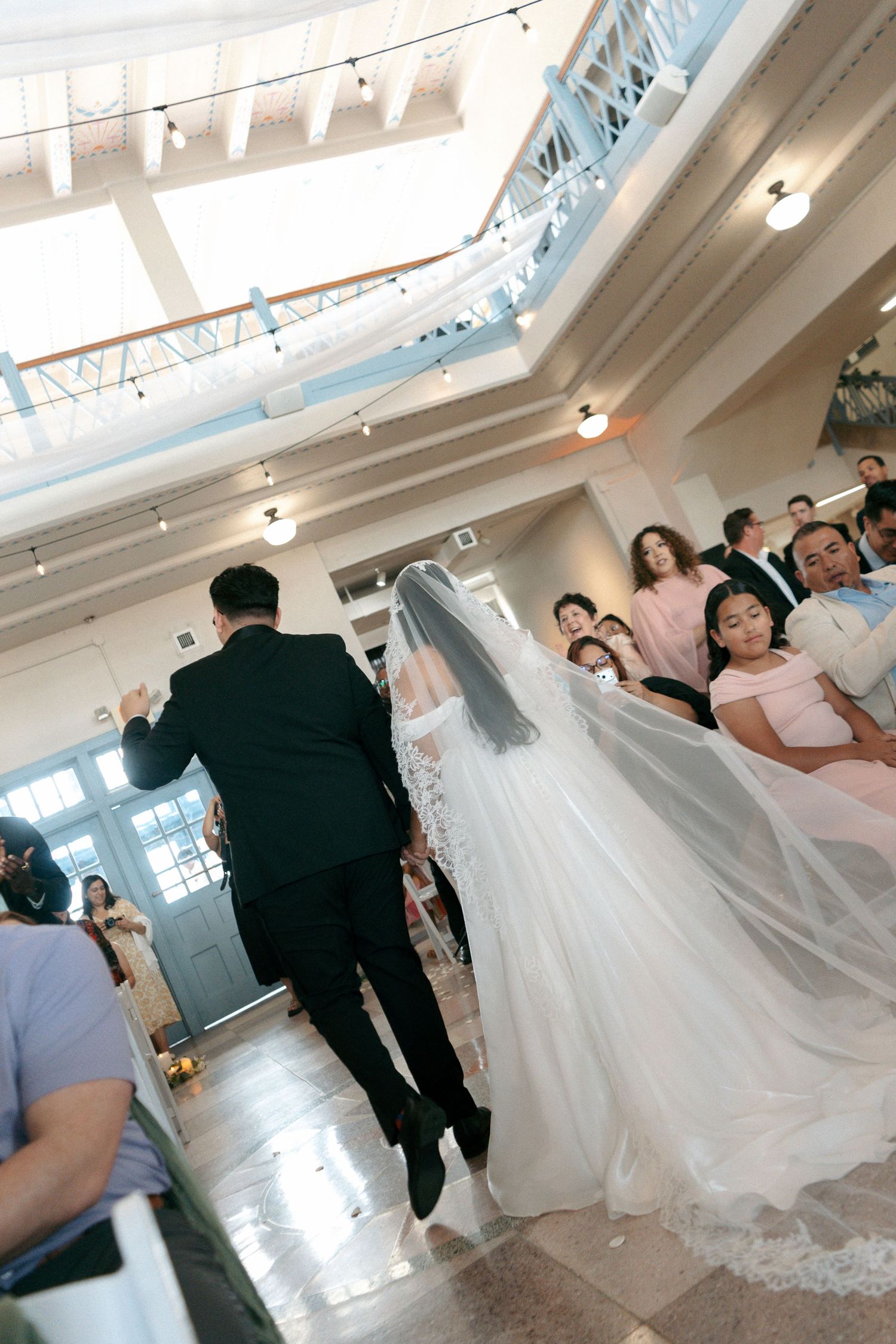 Bride in white gown and veil walks down the aisle with groom in black suit at elegant indoor wedding ceremony.