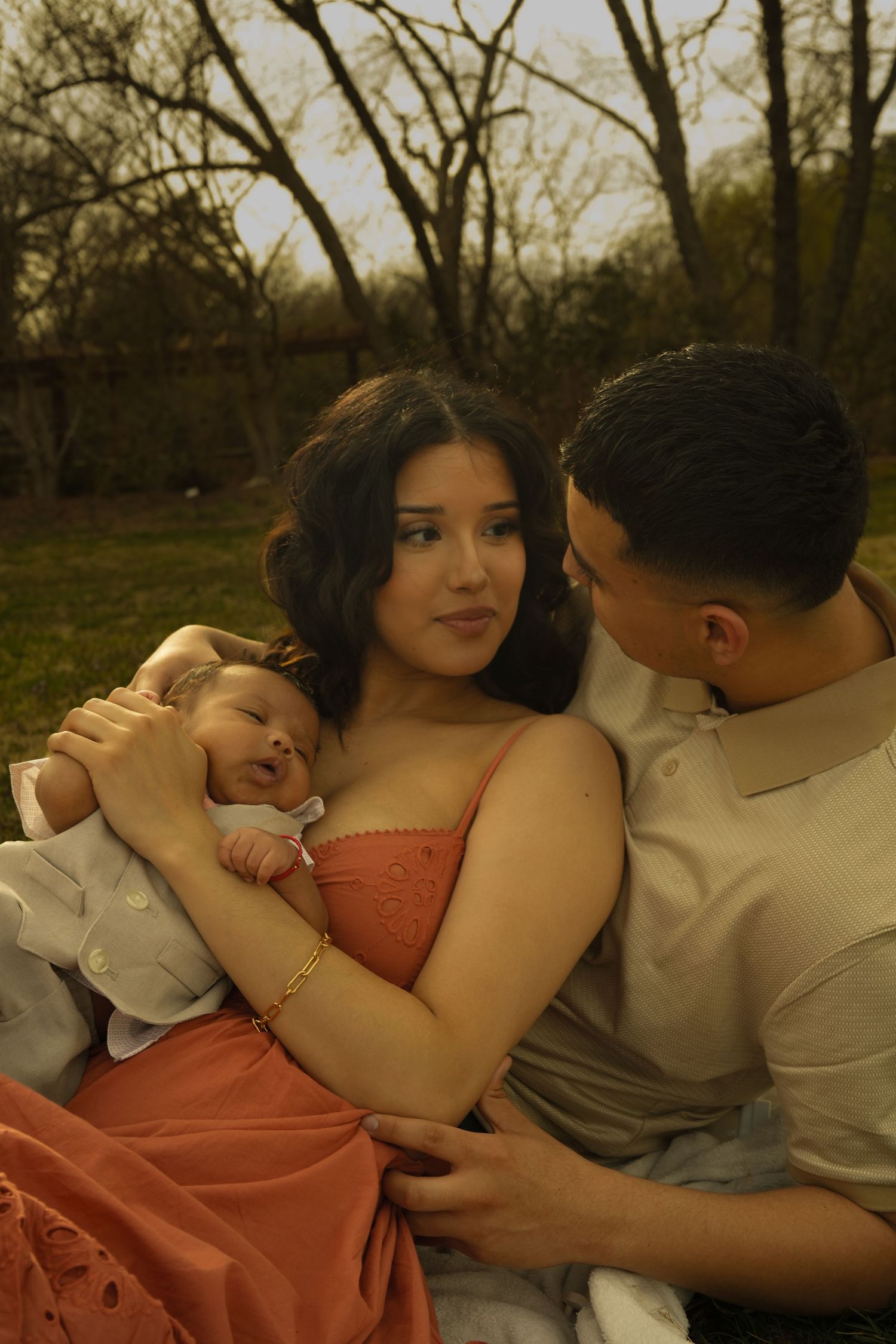 New parents sitting outdoors in evening light, mother in coral dress cradling sleeping newborn baby.