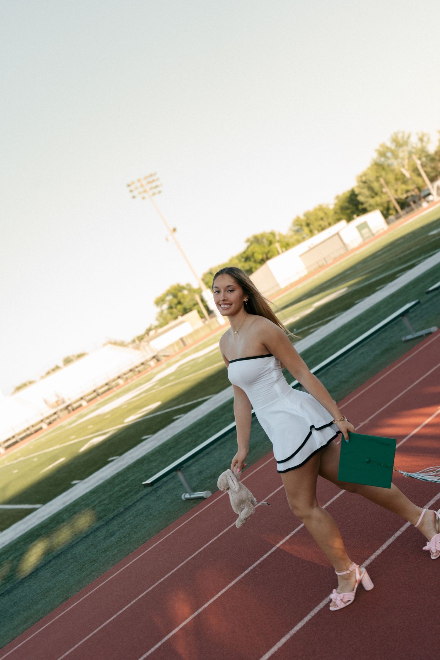 Graduate in white dress carries green cap and heels while walking on track.