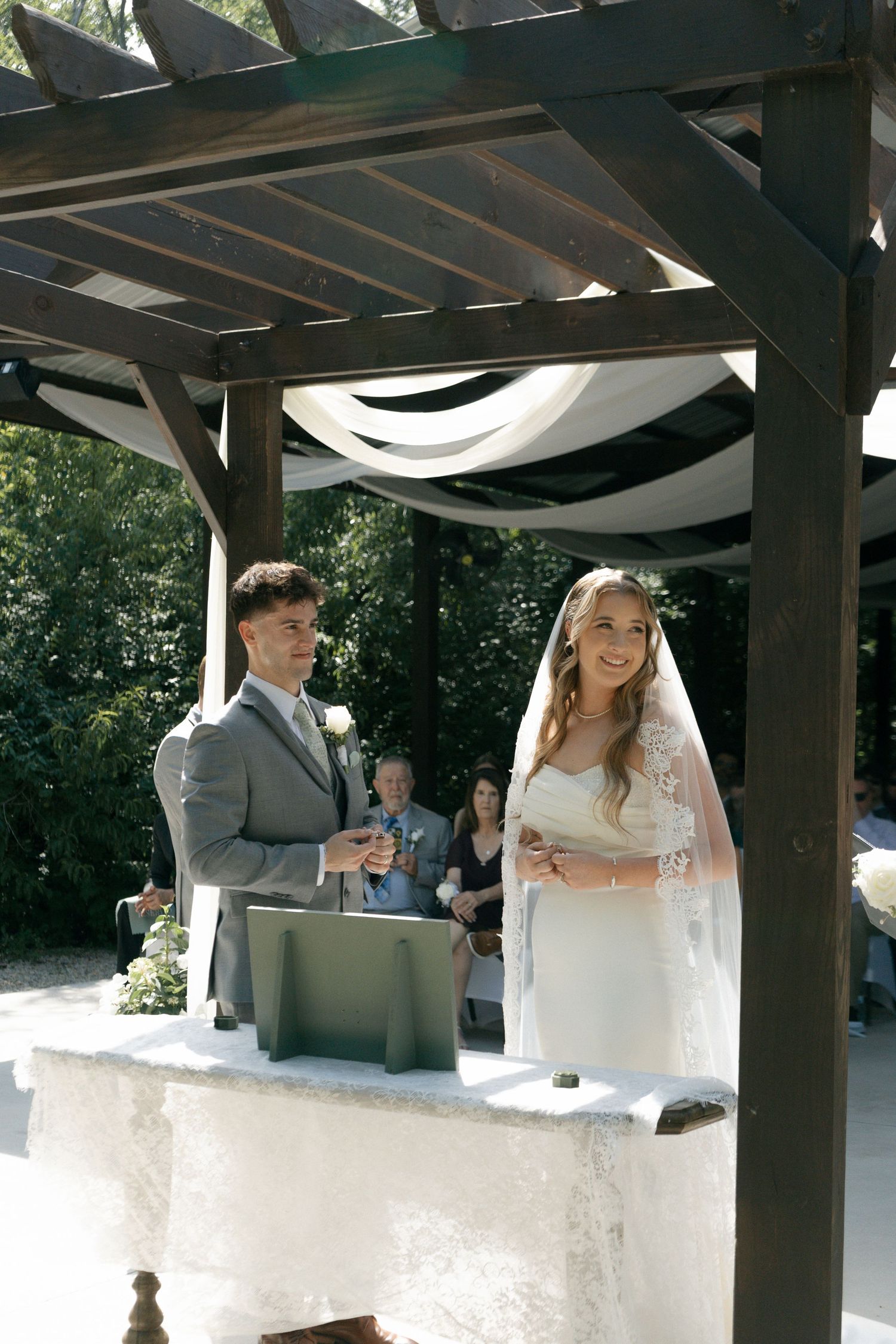 Bride and groom smiling during outdoor wedding ceremony under wooden pergola with white draping.