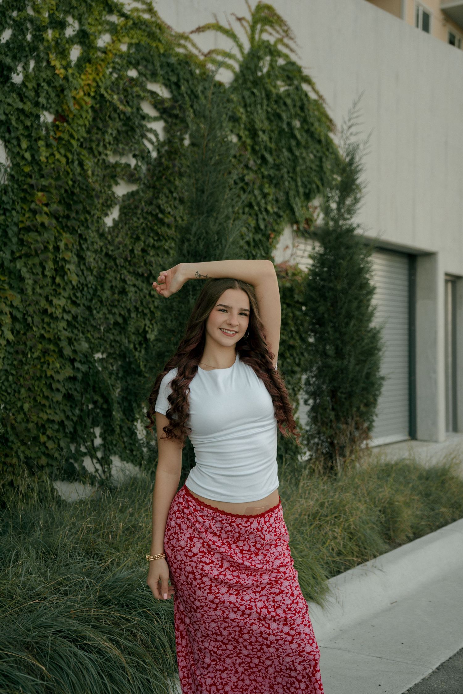Young woman in white tee and red floral skirt posing with arm raised near ivy-covered building.