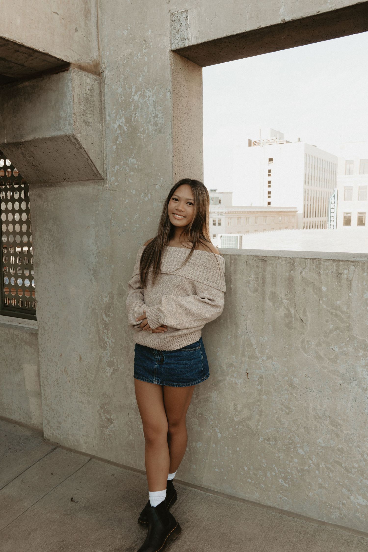 A smiling young woman in a beige sweater and denim mini skirt poses on an urban rooftop.