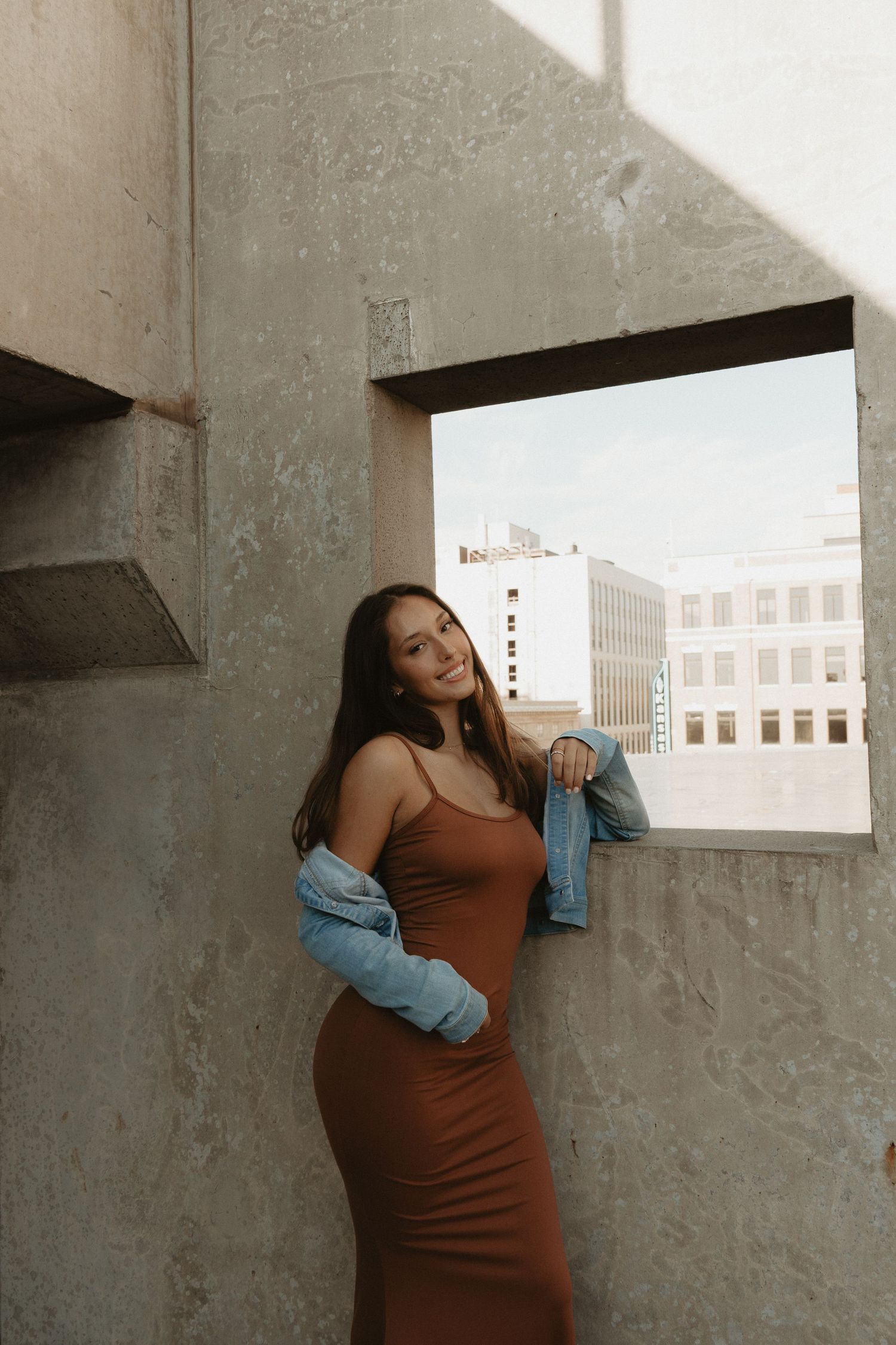 A stylish young woman in a brown dress and denim jacket poses against a concrete rooftop wall.