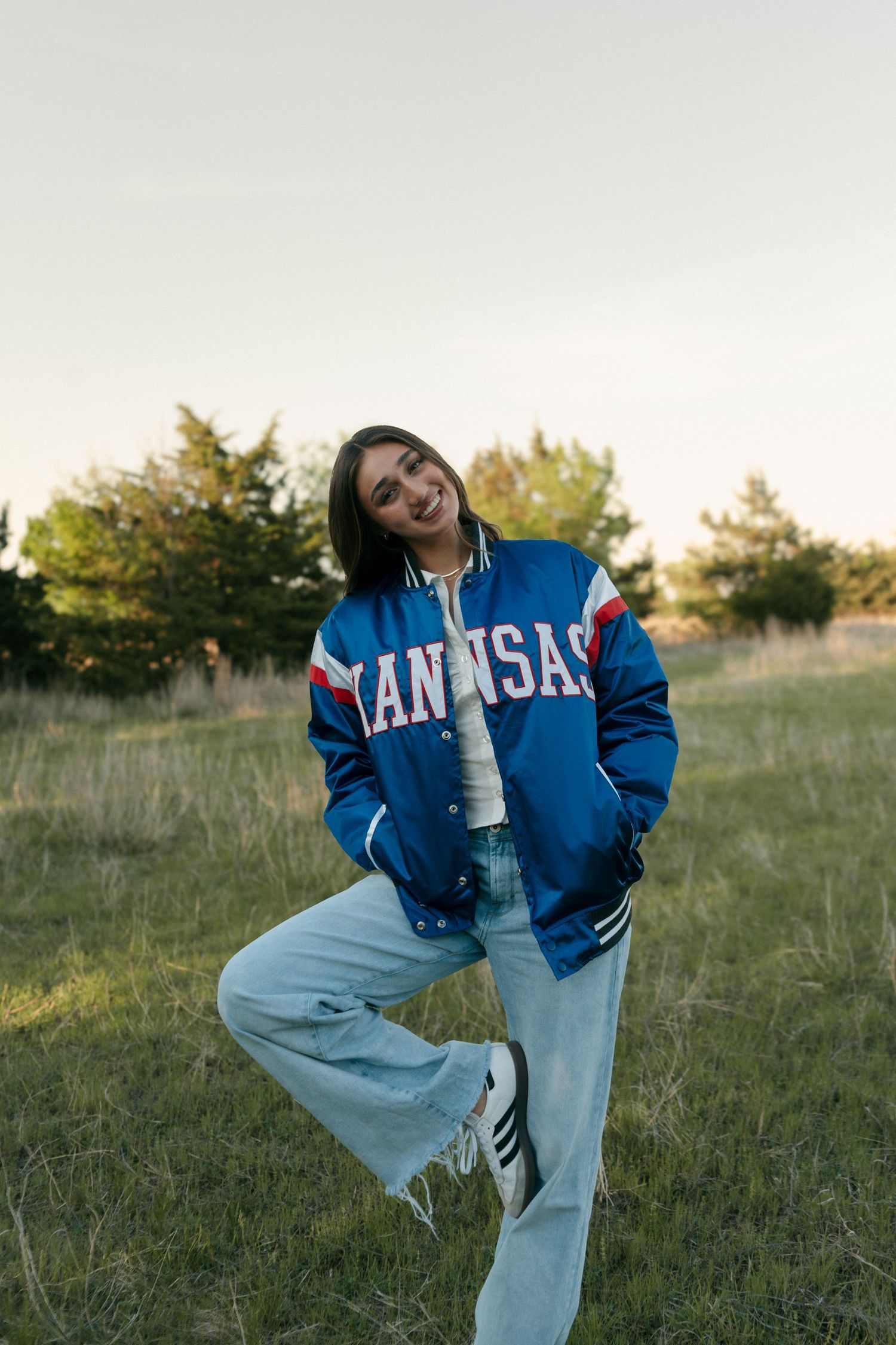 Smiling young woman posing in blue Kansas Jayhawks bomber jacket and light jeans in an open field.