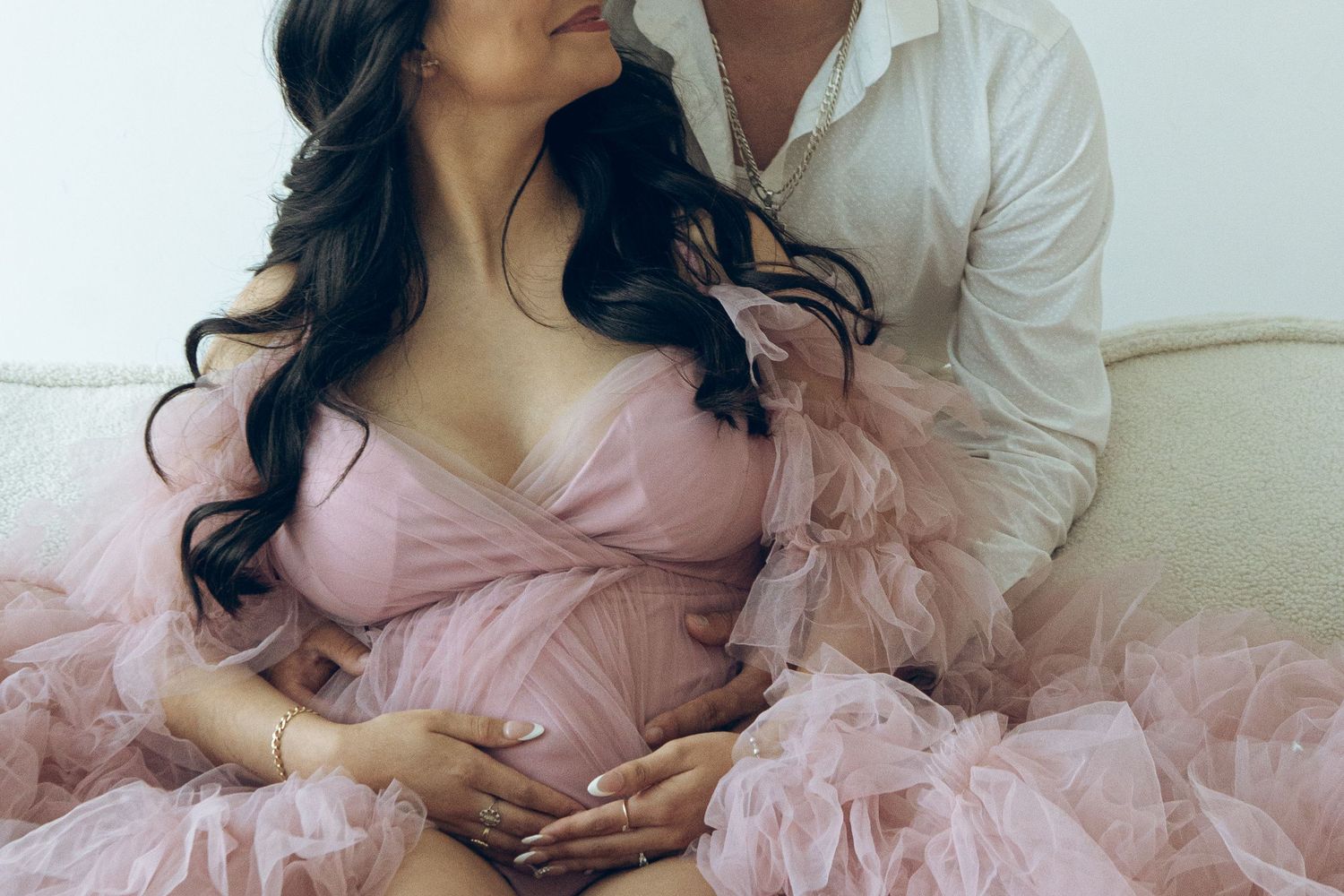 Pregnant woman in pink tulle maternity gown sits with partner on white couch during maternity photoshoot.