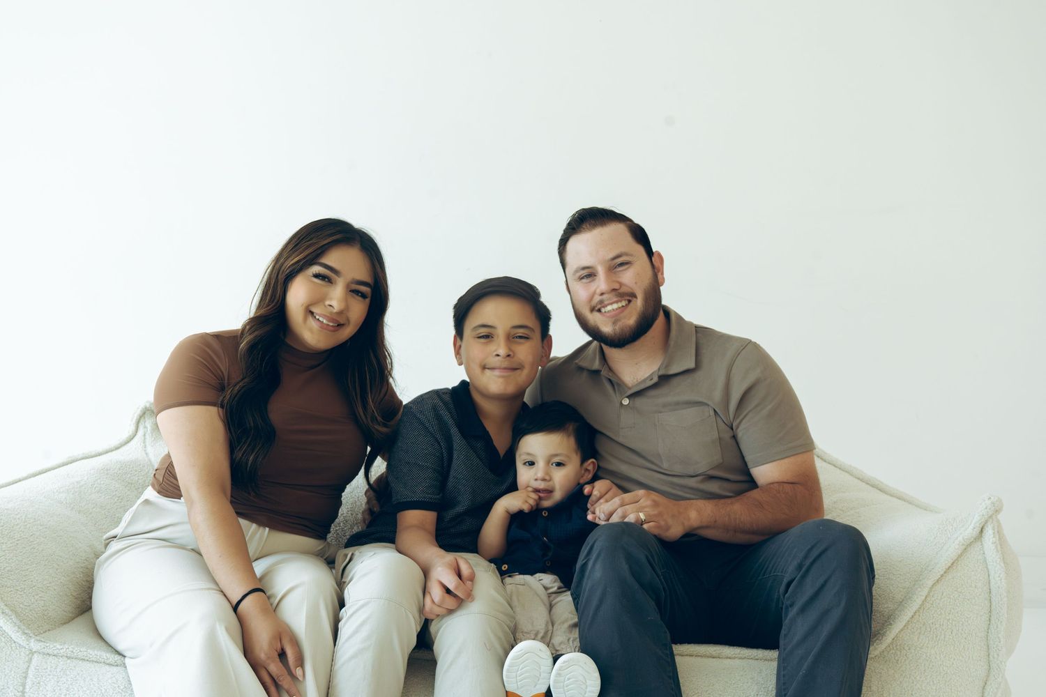 Smiling family of four with two boys sitting together on a white studio couch.