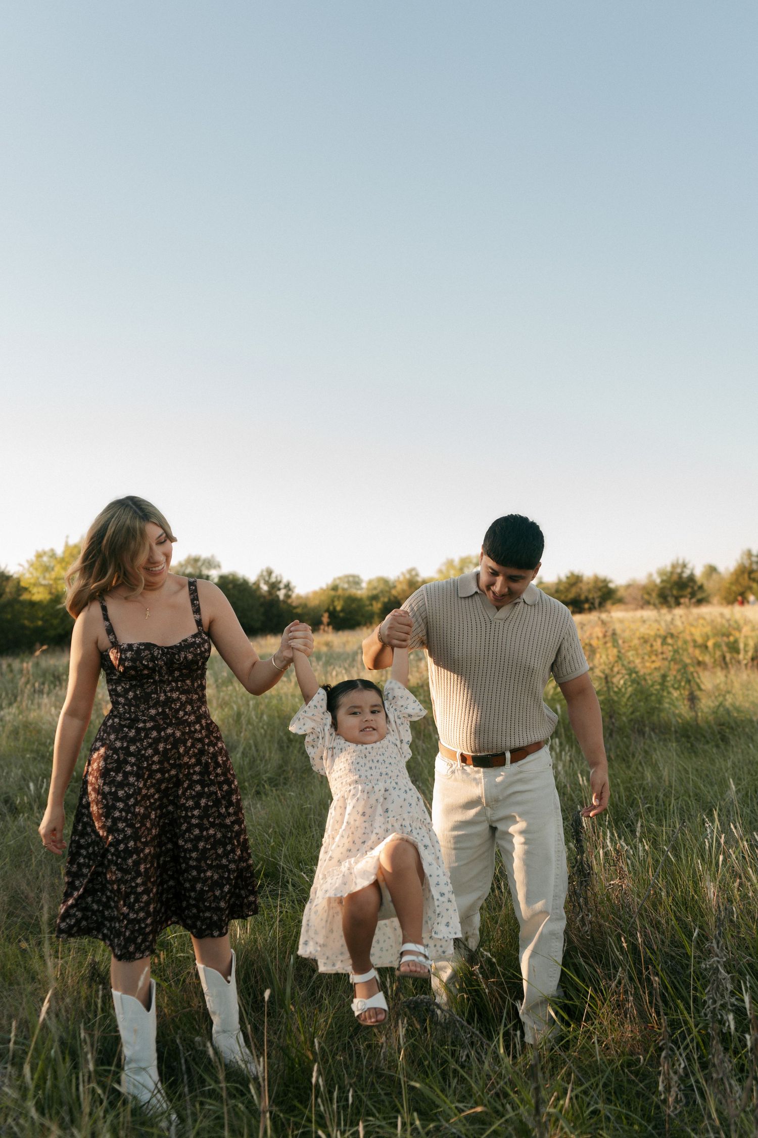 A young family swings their toddler daughter between them while walking through a sunny green meadow at golden hour.