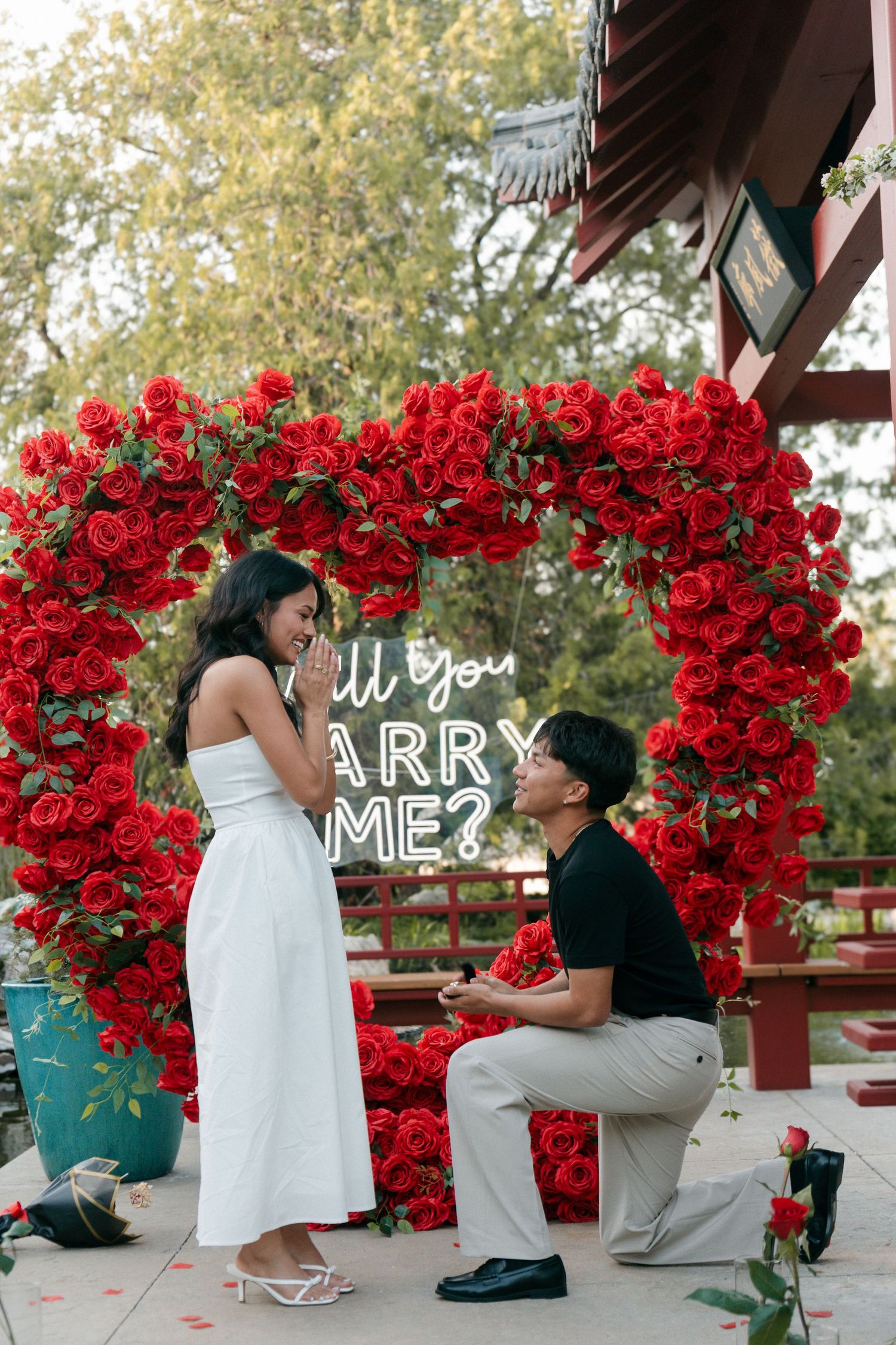 Man kneeling on one knee proposing to woman under red rose arch with Will You Marry Me neon sign.