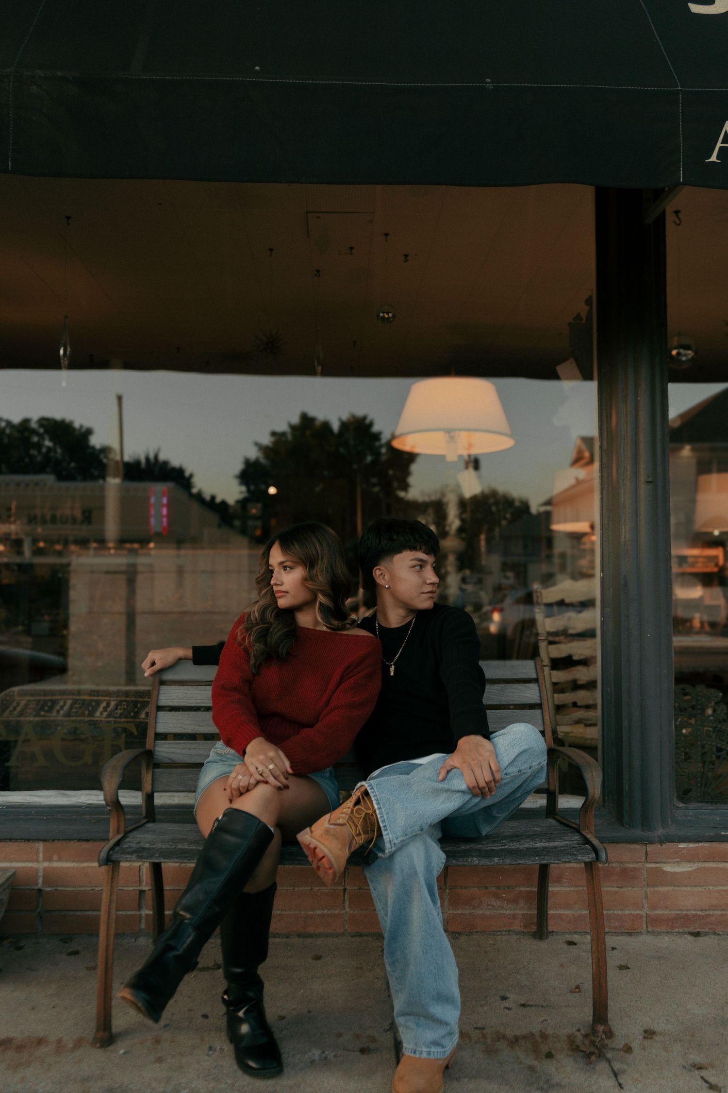 Couple sitting close together on a bench outside a shop at dusk, woman in red sweater and black boots.