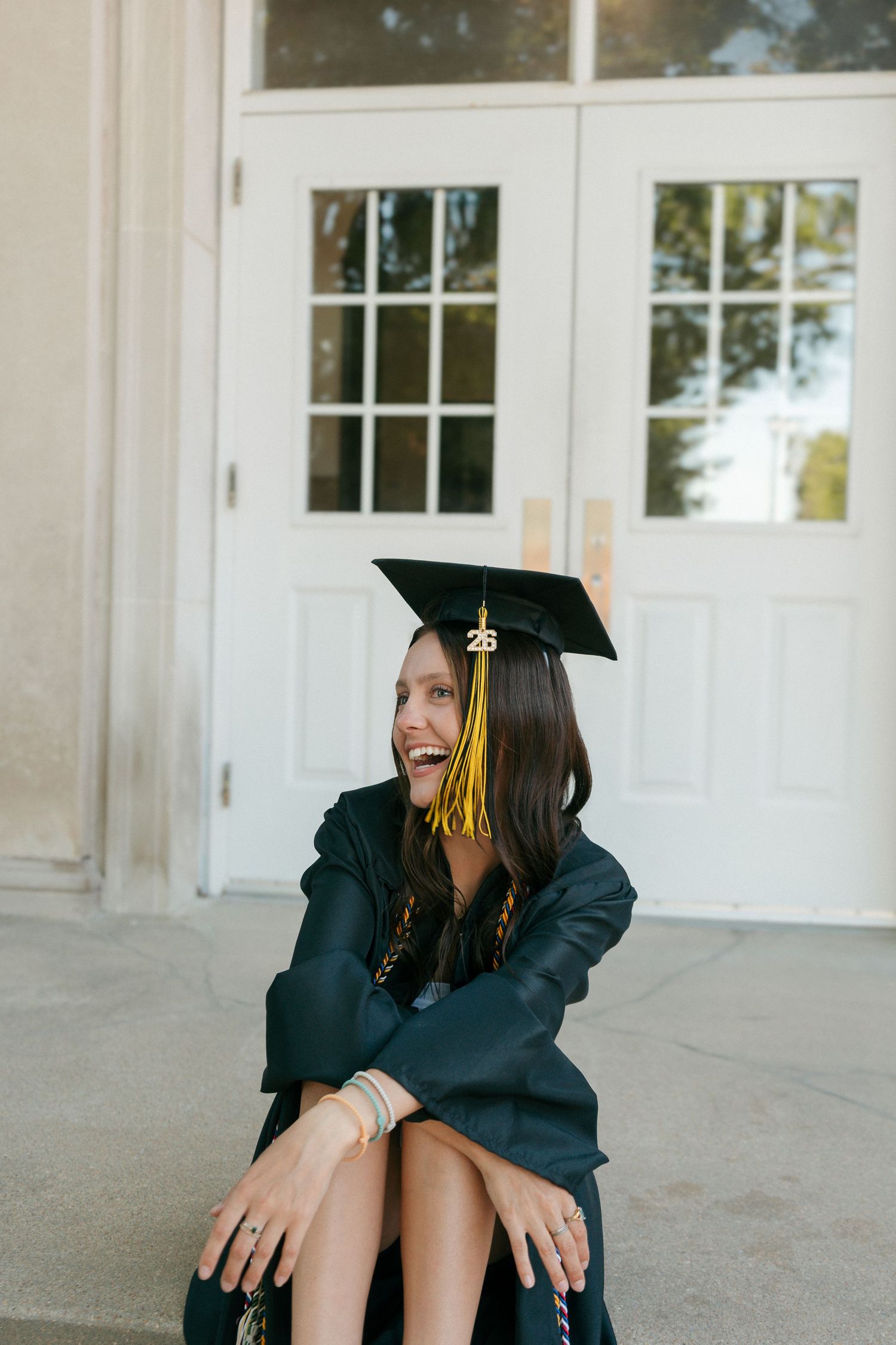 A smiling young woman in a green graduation cap and gown sits outdoors in front of white double doors.