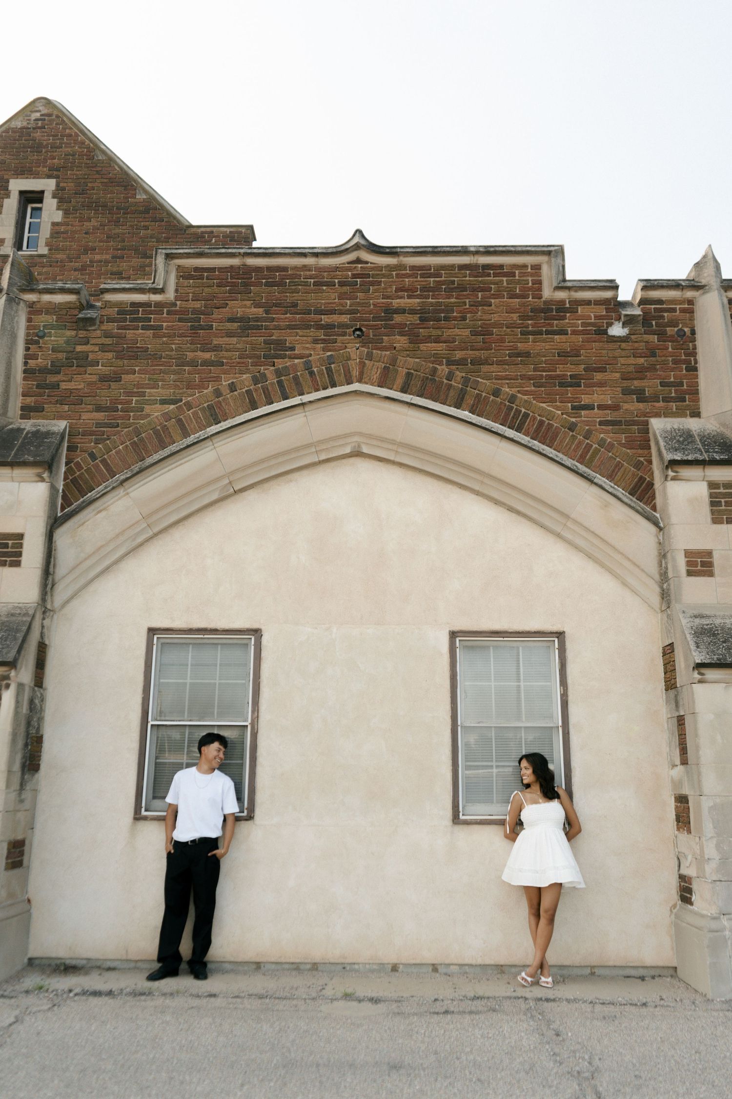 Couple in matching white outfits stand apart against a cream and brick building wall.