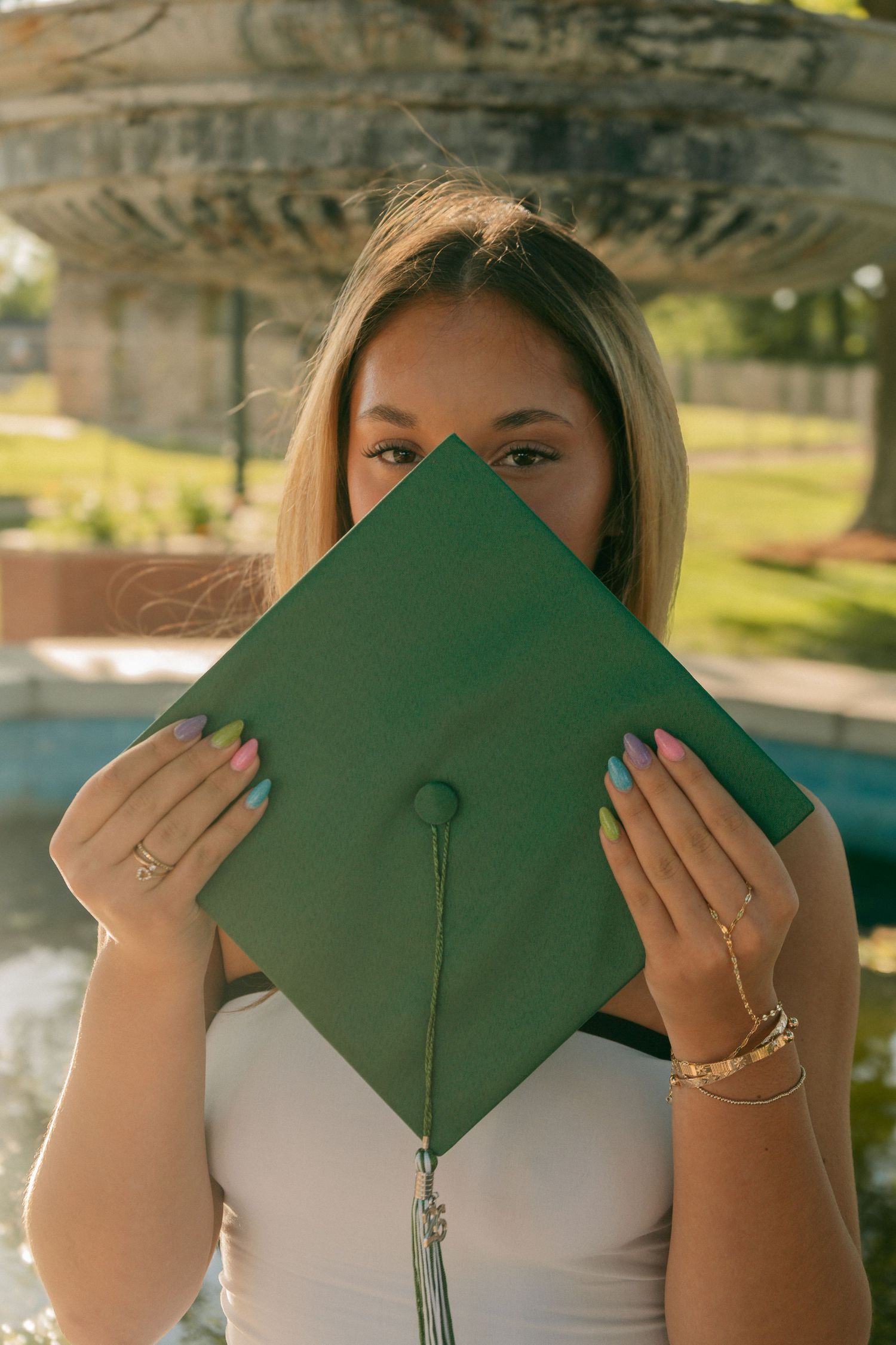 Graduate hiding face behind green mortarboard cap near outdoor fountain.