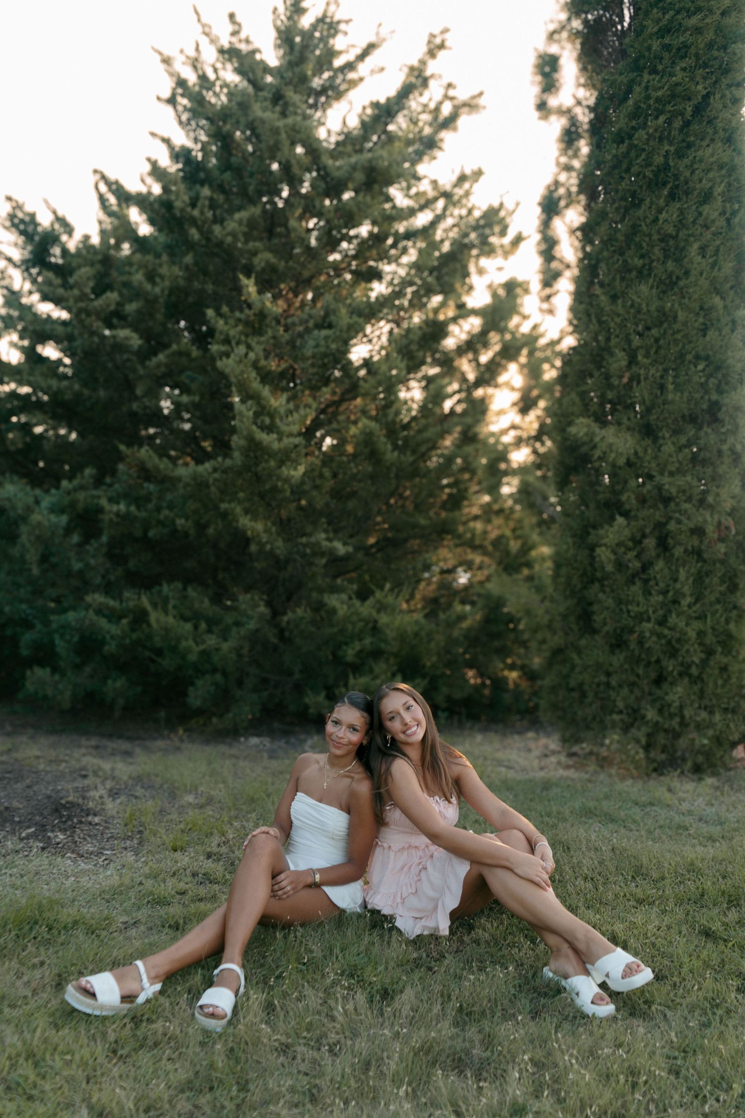 Two young women in white and pink dresses sitting back-to-back on grass surrounded by tall evergreen trees.