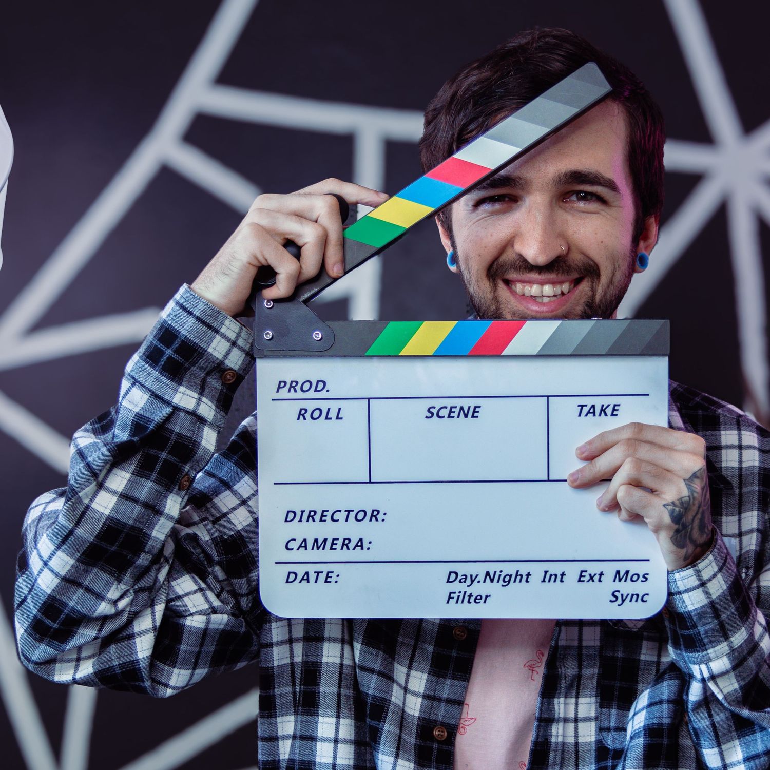 Smiling man holding a film clapperboard in front of his face, wearing a plaid shirt against a dark background.