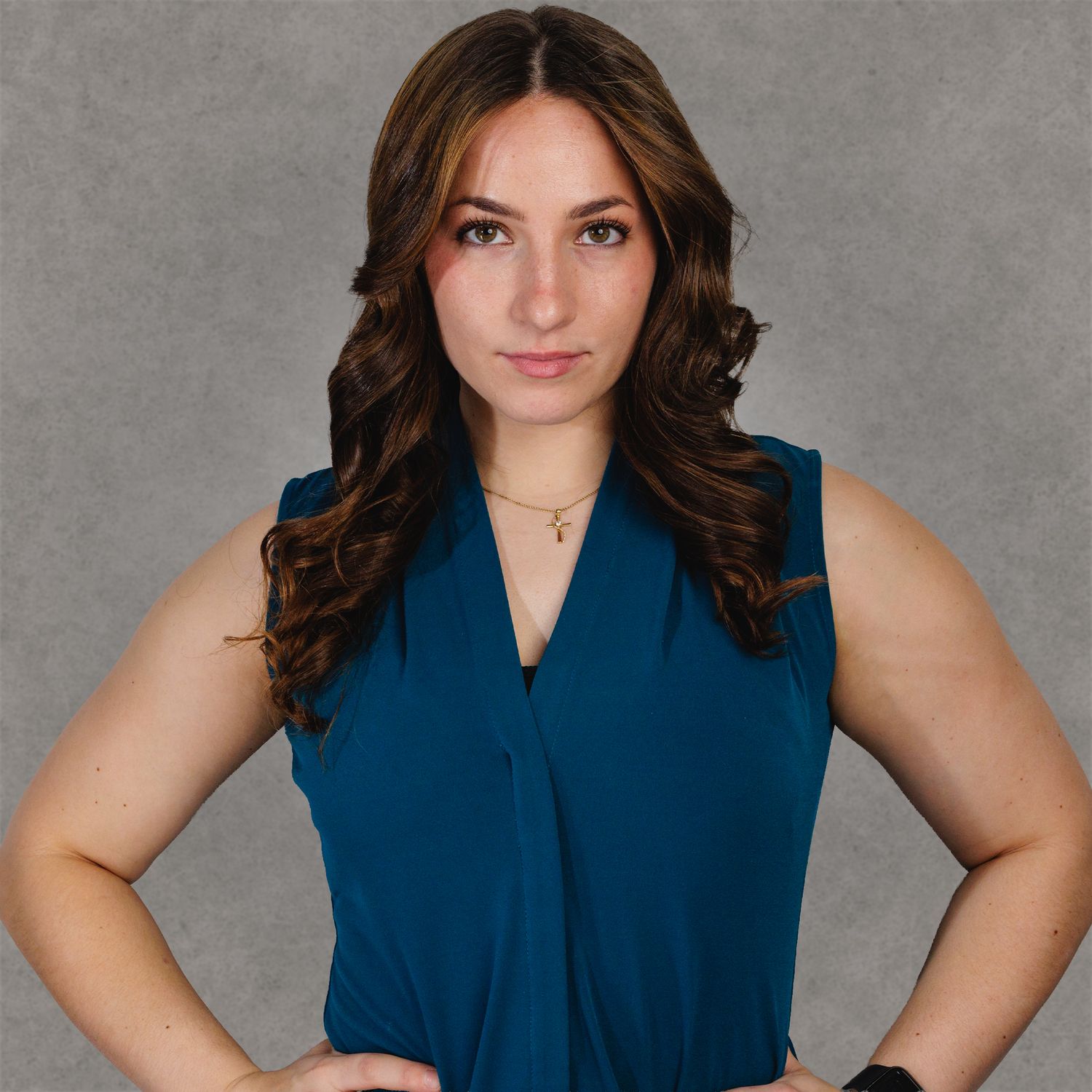 Professional headshot of a young woman in a teal sleeveless top, with long wavy brown hair, hands on hips.